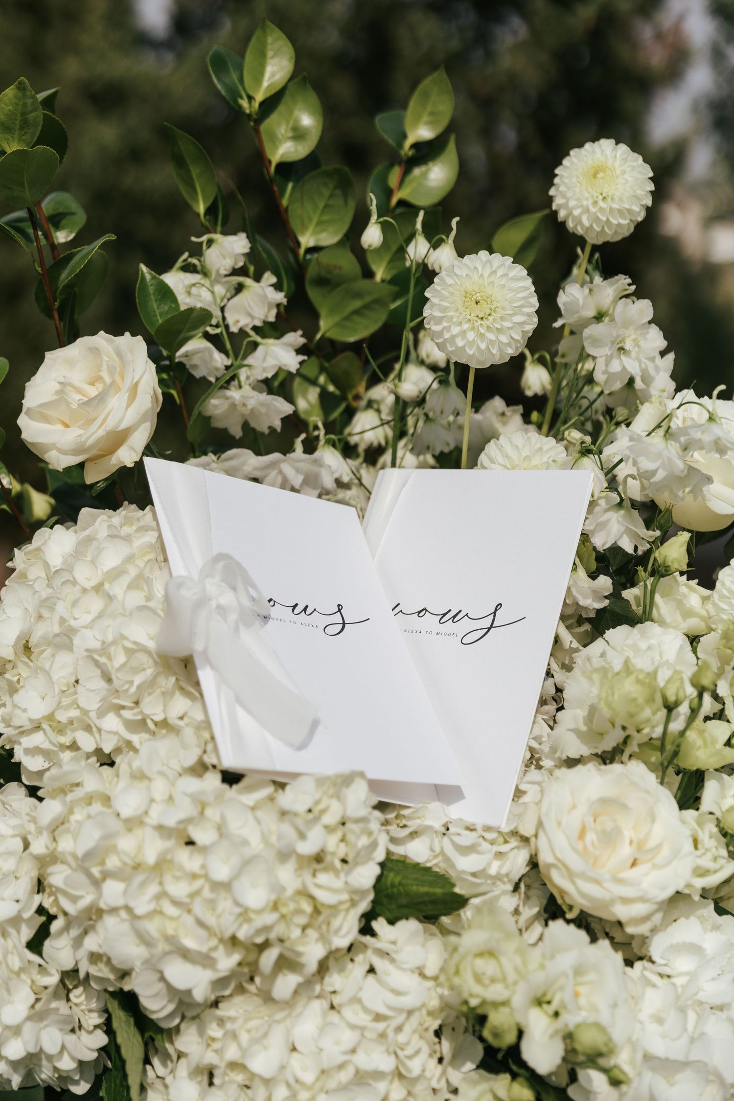 White roses and hydrangeas frame a handwritten wedding note against green foliage.