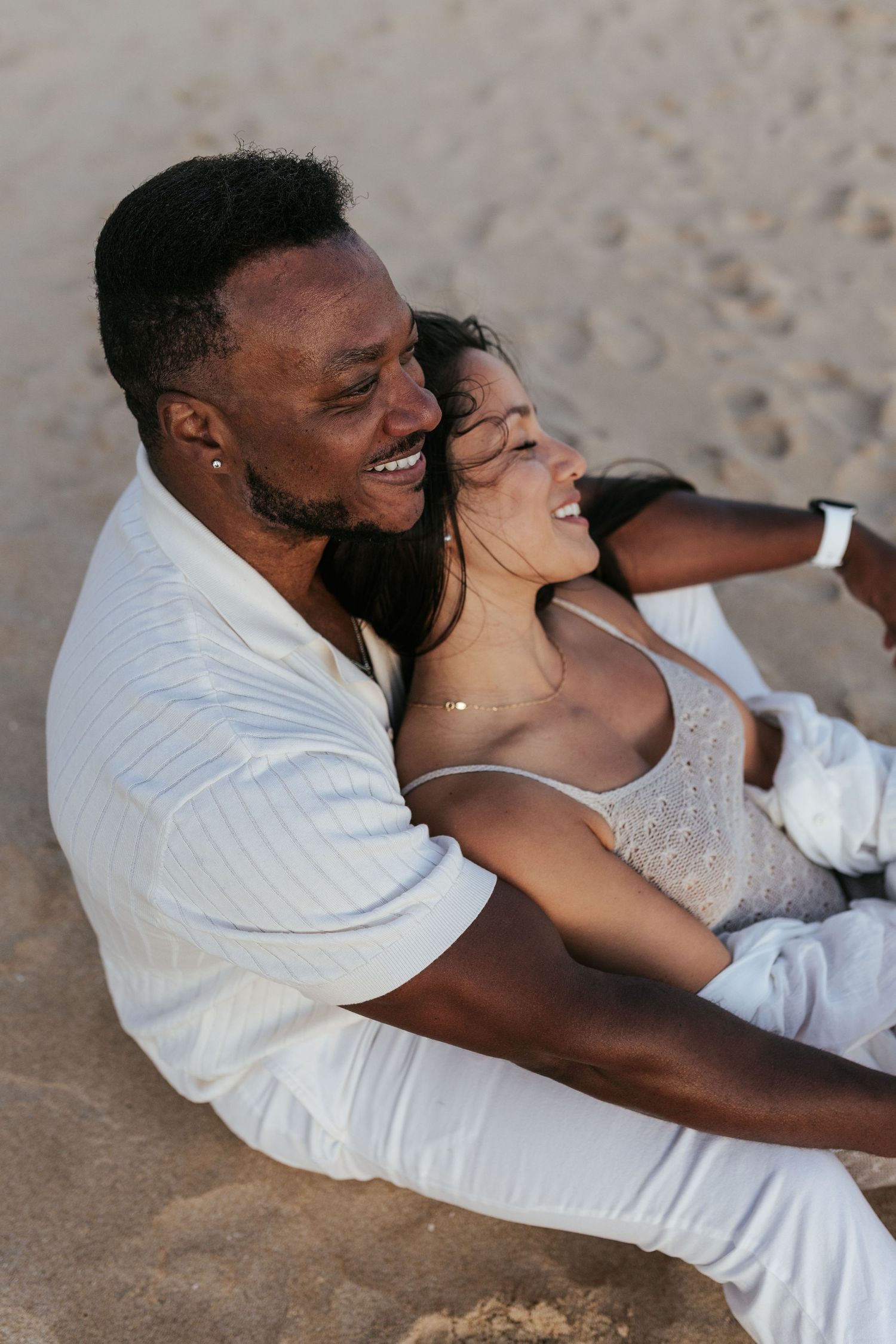 A joyful couple wearing white clothing shares an intimate moment while sitting on a sandy beach.