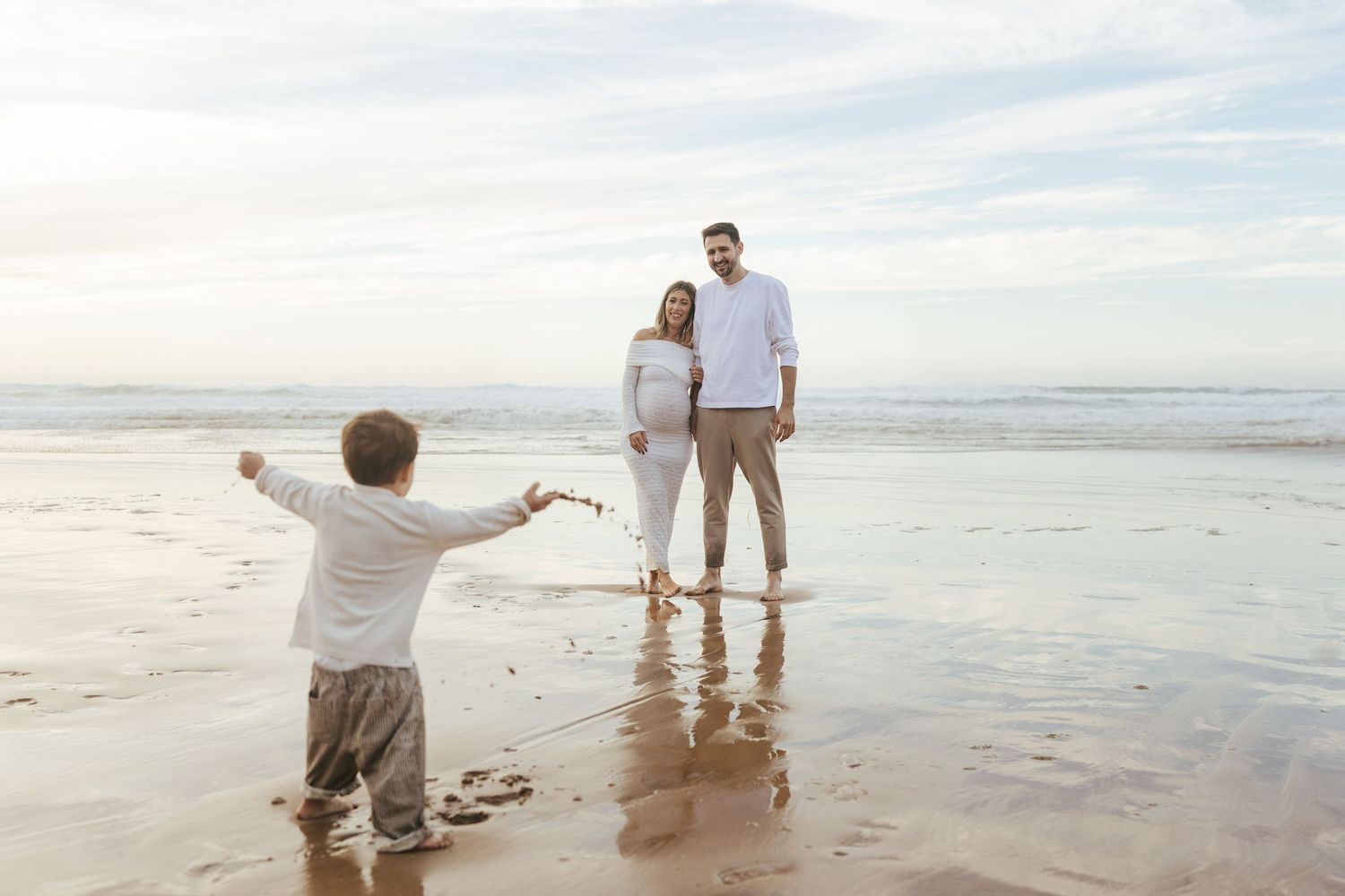 A couple walks hand in hand along a serene beach shoreline at sunset while someone photographs them from behind.