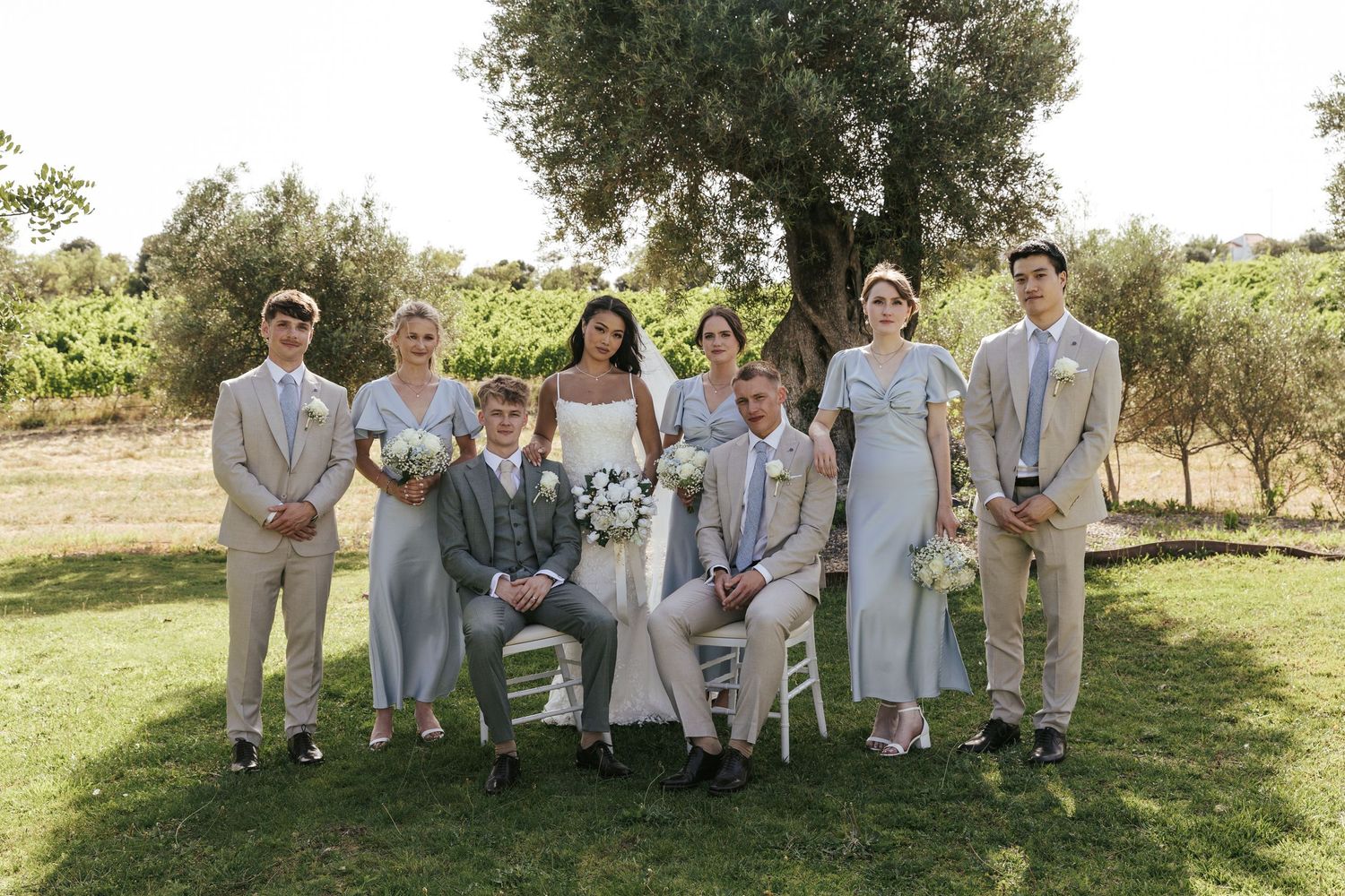Wedding party poses outdoors in a vineyard, wearing neutral suits and sage bridesmaid dresses with bride and groom seated center.