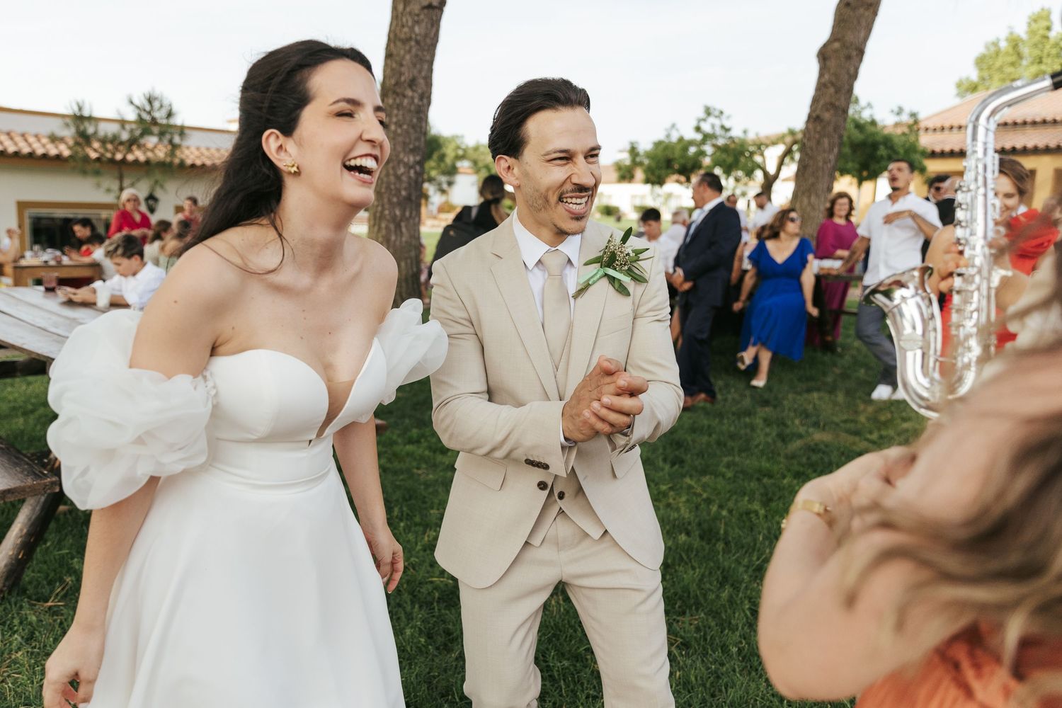 Joyful couple and wedding party dance together on a grassy lawn during an outdoor celebration.