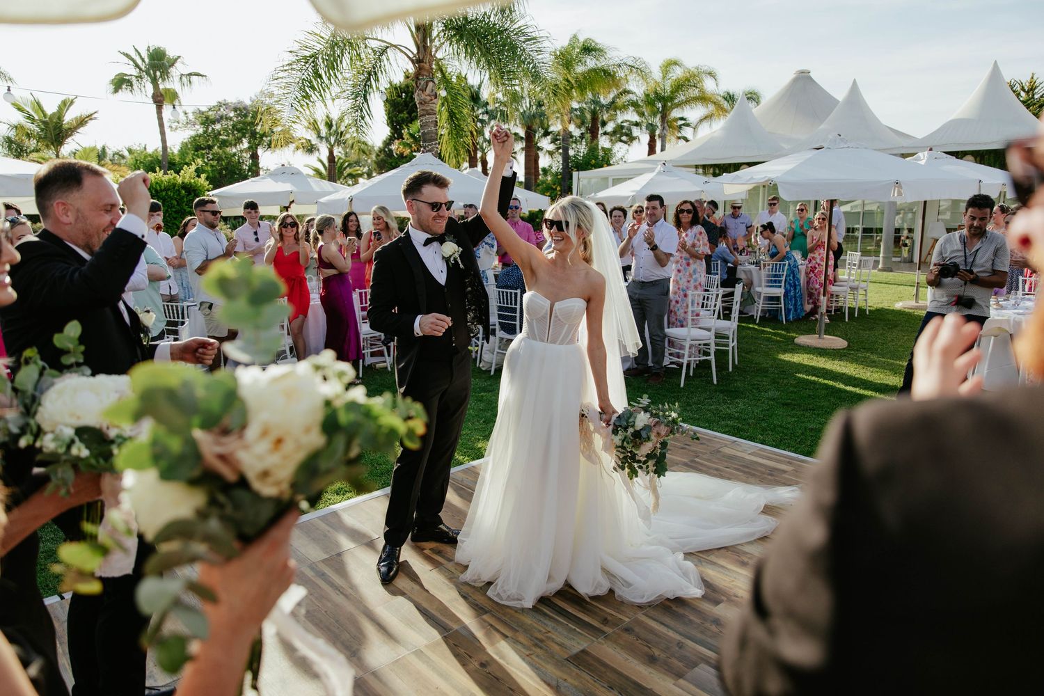 A wedding reception celebration outdoors with white tents, palm trees, and guests mingling on a sunny day.