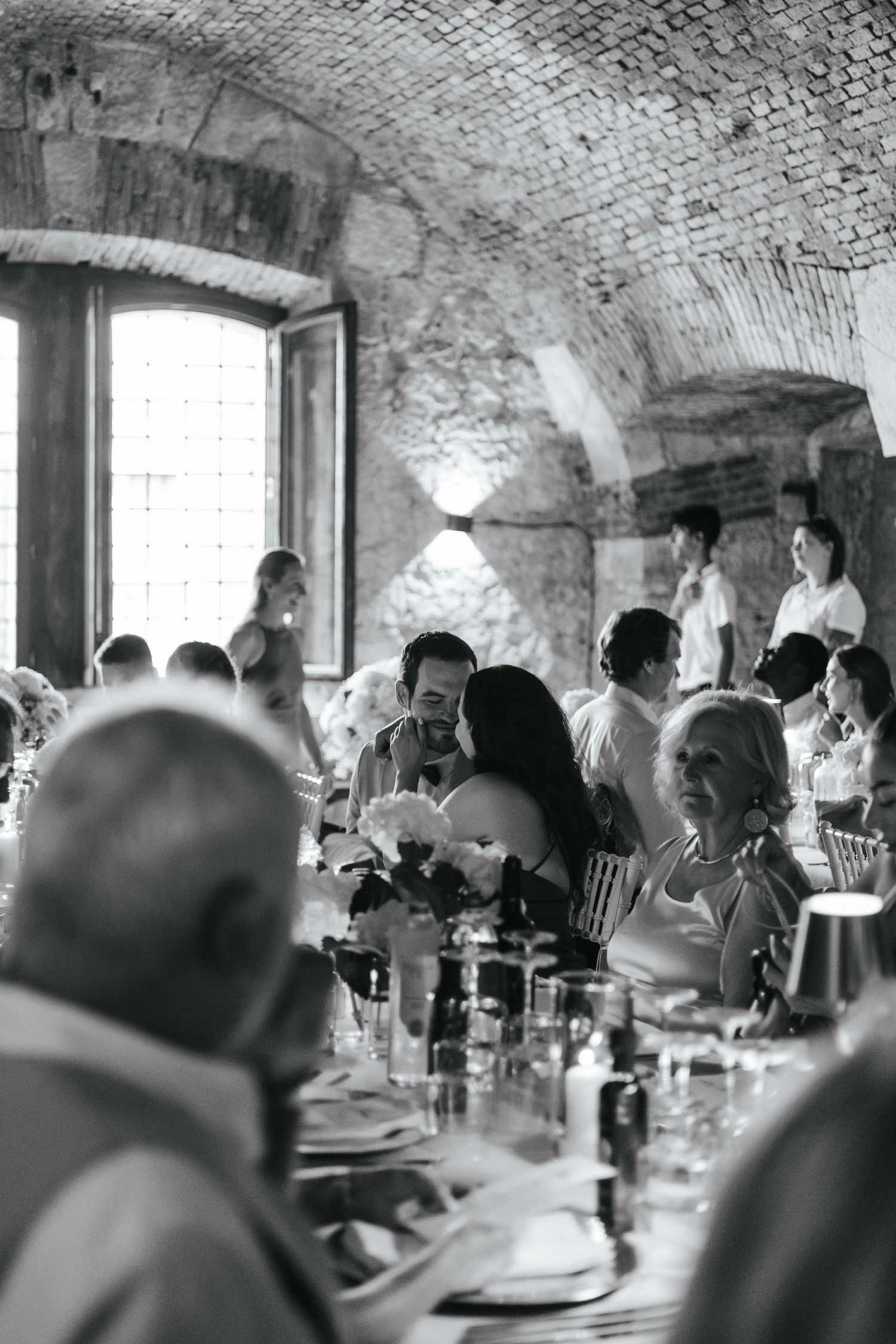 Wedding guests enjoy dinner and conversation in a rustic stone-walled venue with arched ceilings.