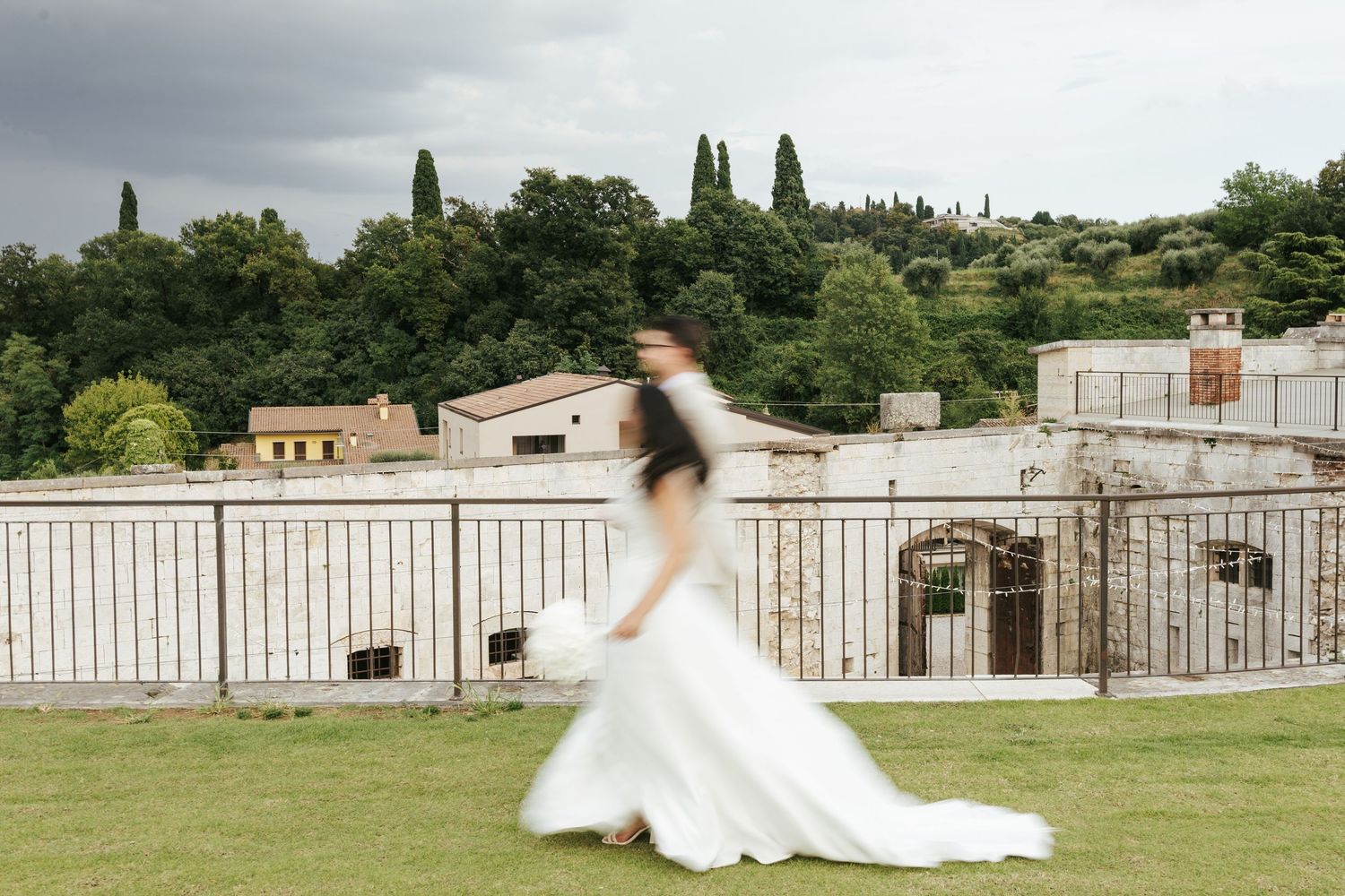 Bride in flowing white dress twirling on terrace with Italian countryside in background.