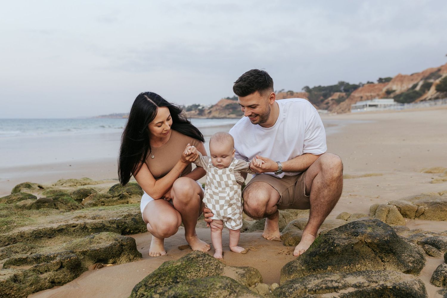 A family spends time together on a rocky beach with cliffs and ocean waves in the background.