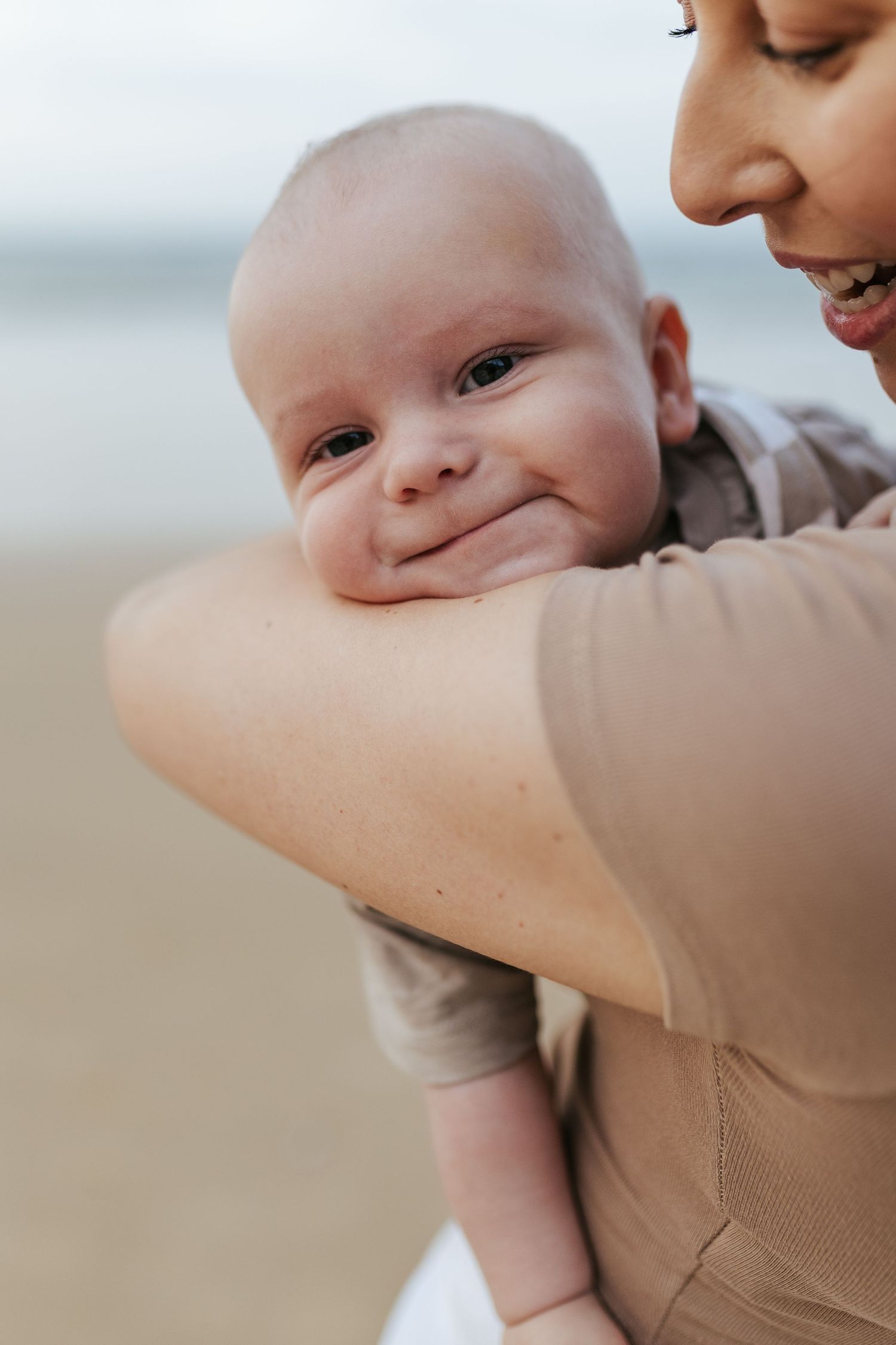 Close-up portrait of a smiling baby being held at the beach, showing rosy cheeks and bright expression.