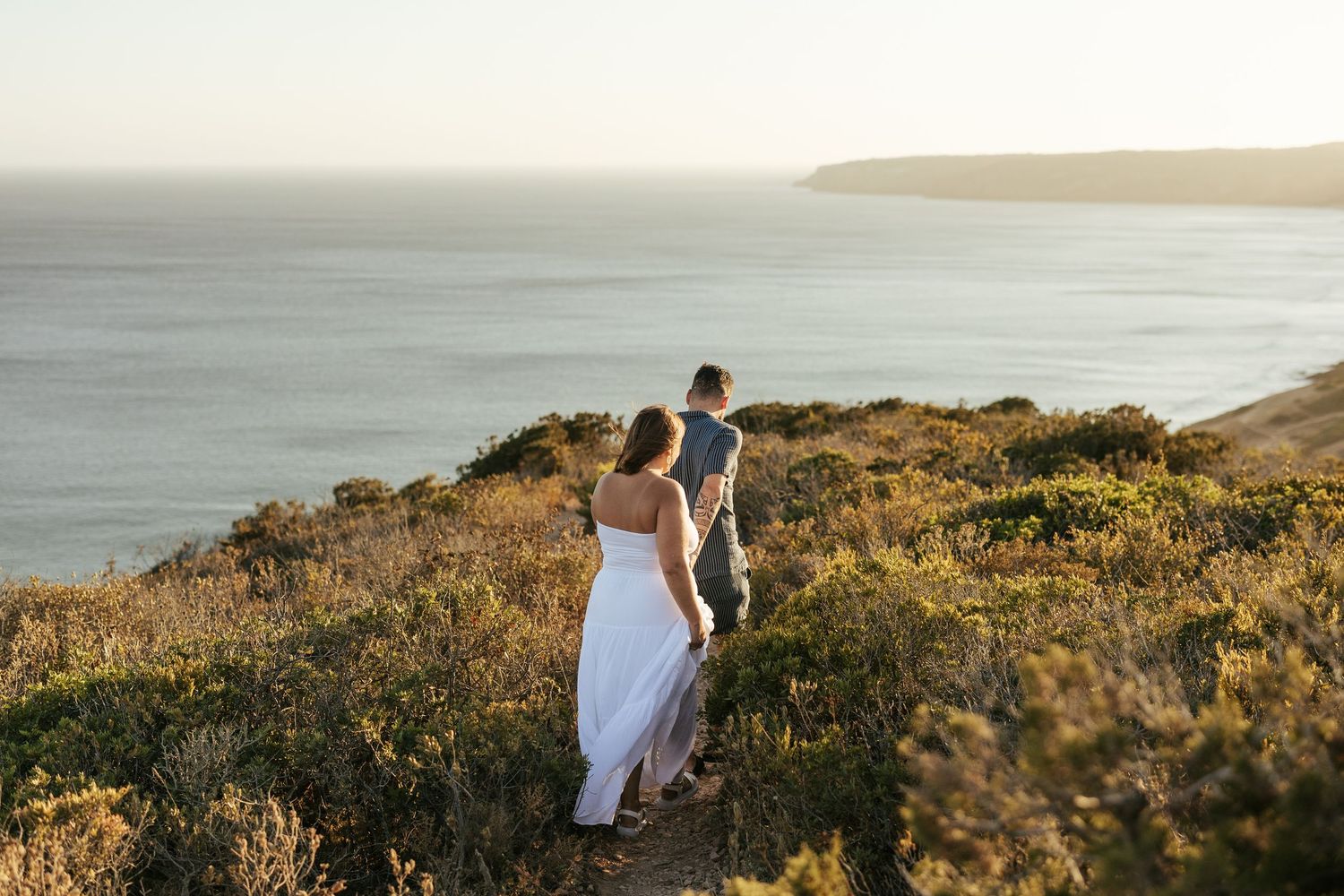 Couple walking on coastal hillside path during golden hour with ocean and distant cliffs in background.