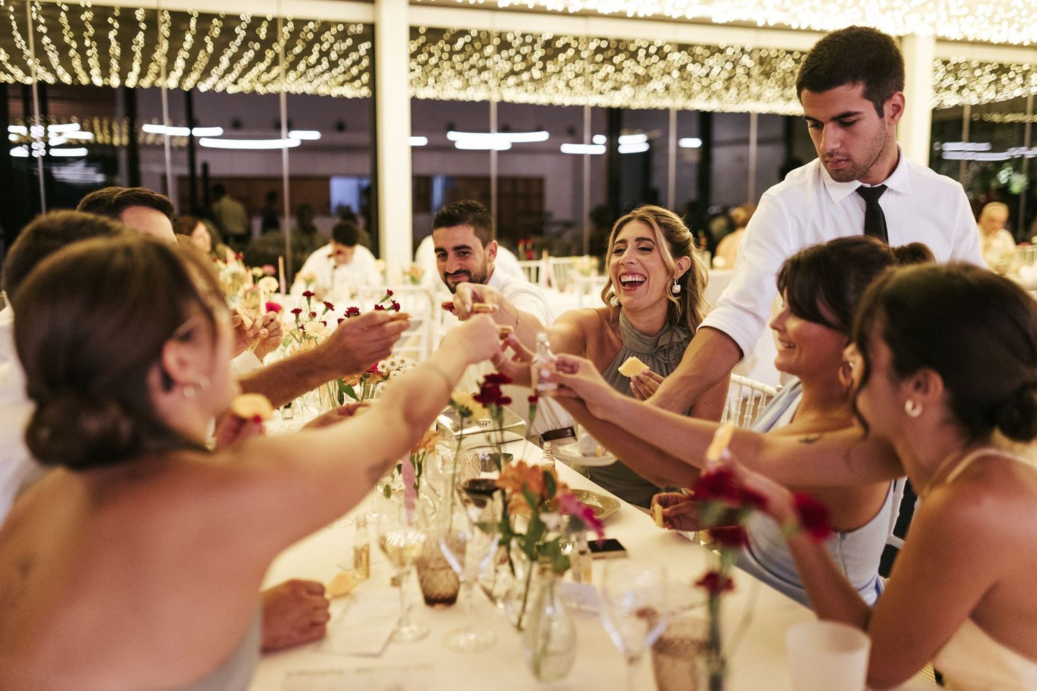 Wedding guests raise glasses in a celebratory toast under a canopy of string lights.