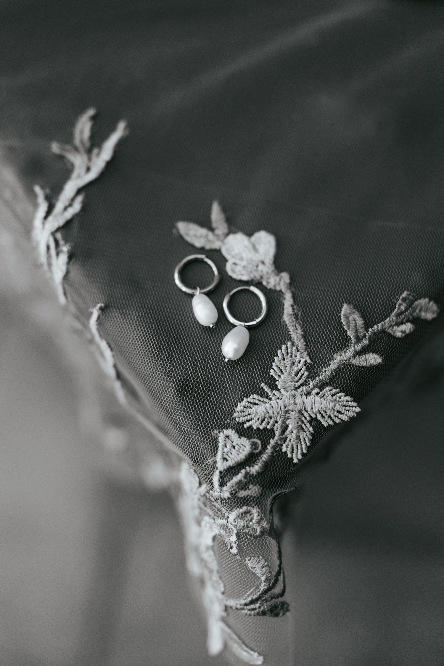 Black and white photo of pearl drop earrings and wedding rings on delicate lace bridal veil with floral embroidery.