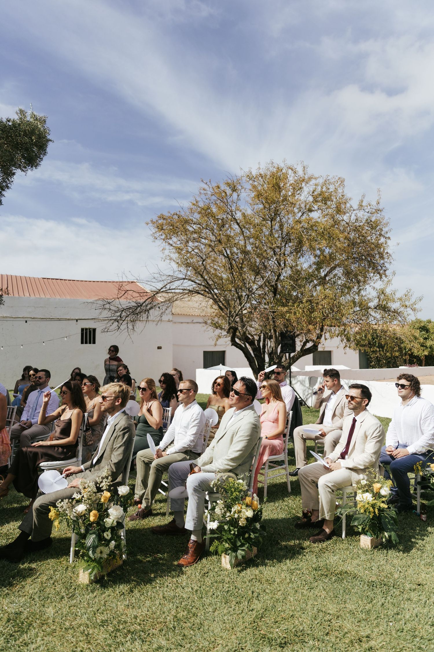 Guests seated outdoors at a sunny wedding ceremony on green lawn, with floral arrangements and a tree in the background.