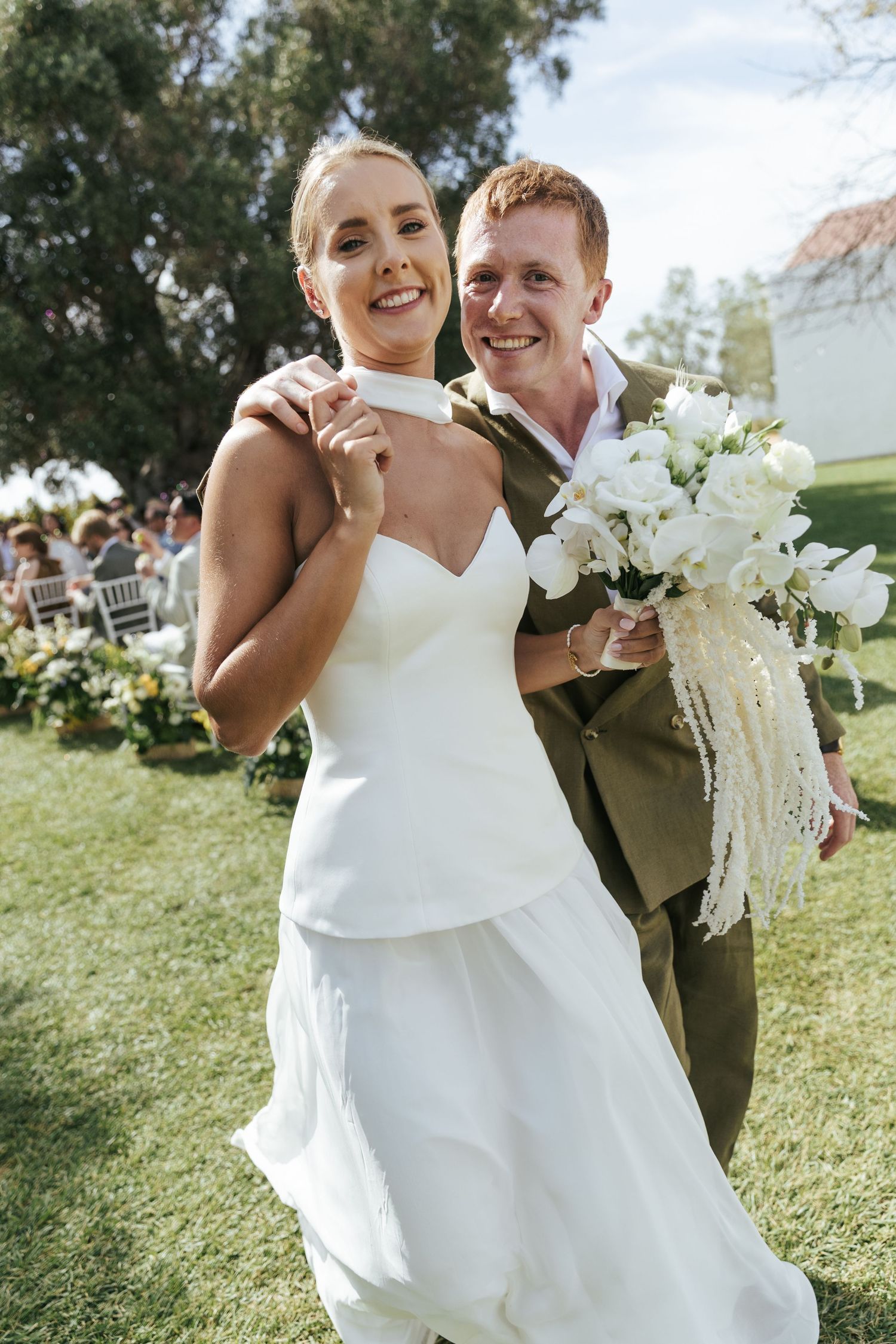 Smiling bride in white gown and groom in olive suit holding white floral bouquet at outdoor wedding.
