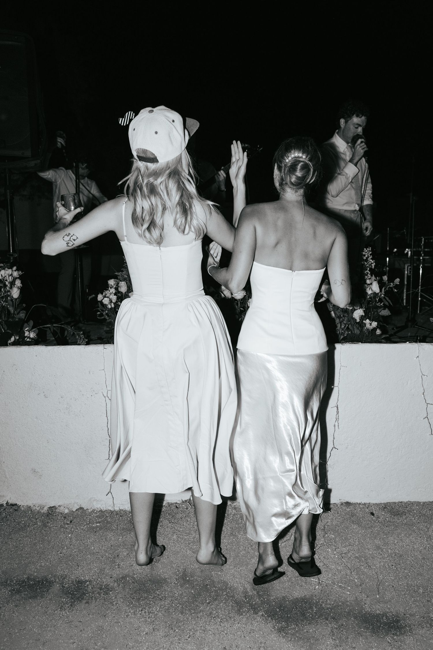 Two women in white dresses dance with raised hands at an outdoor wedding reception.