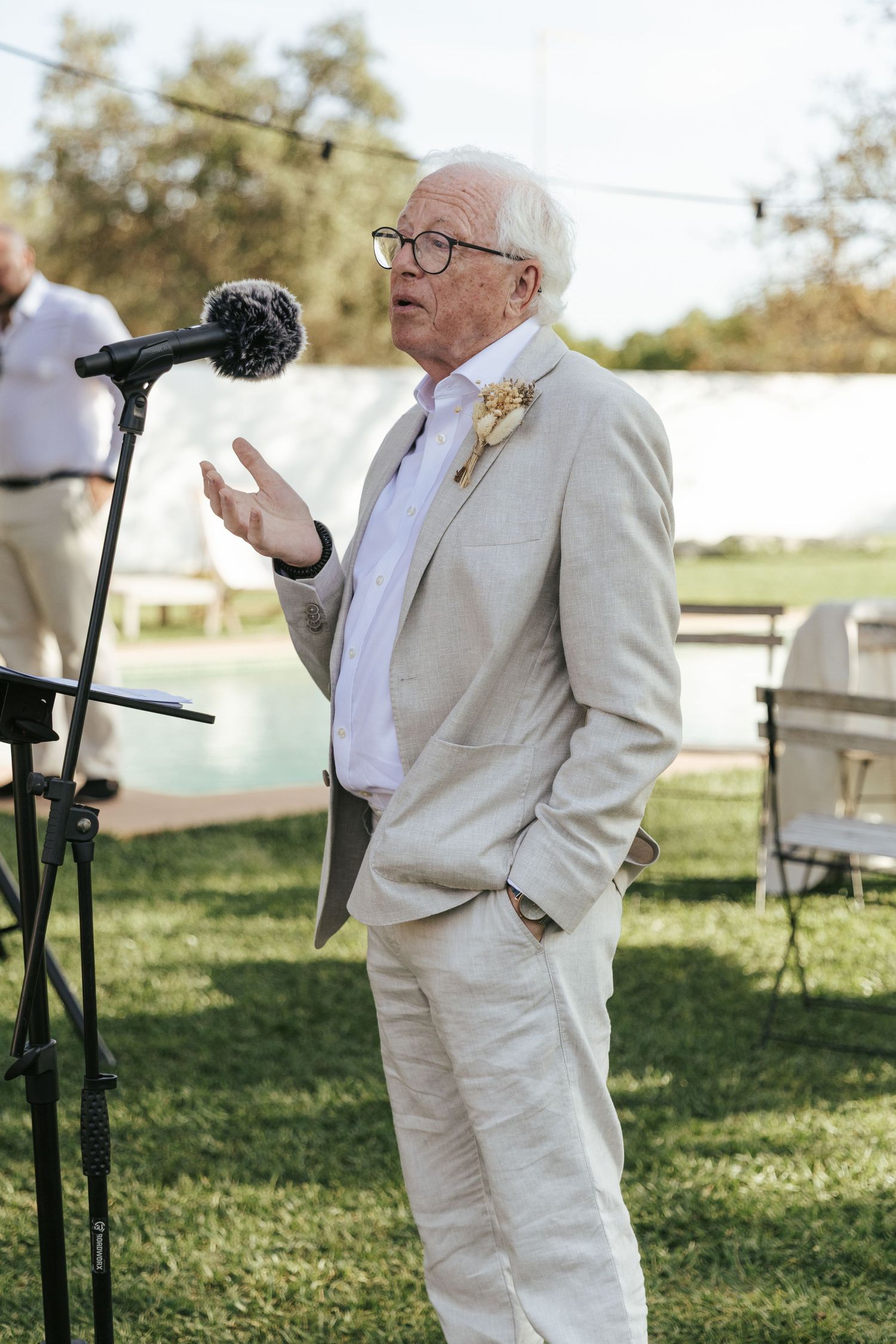 Elderly man in cream linen suit delivering a wedding speech at an outdoor microphone.