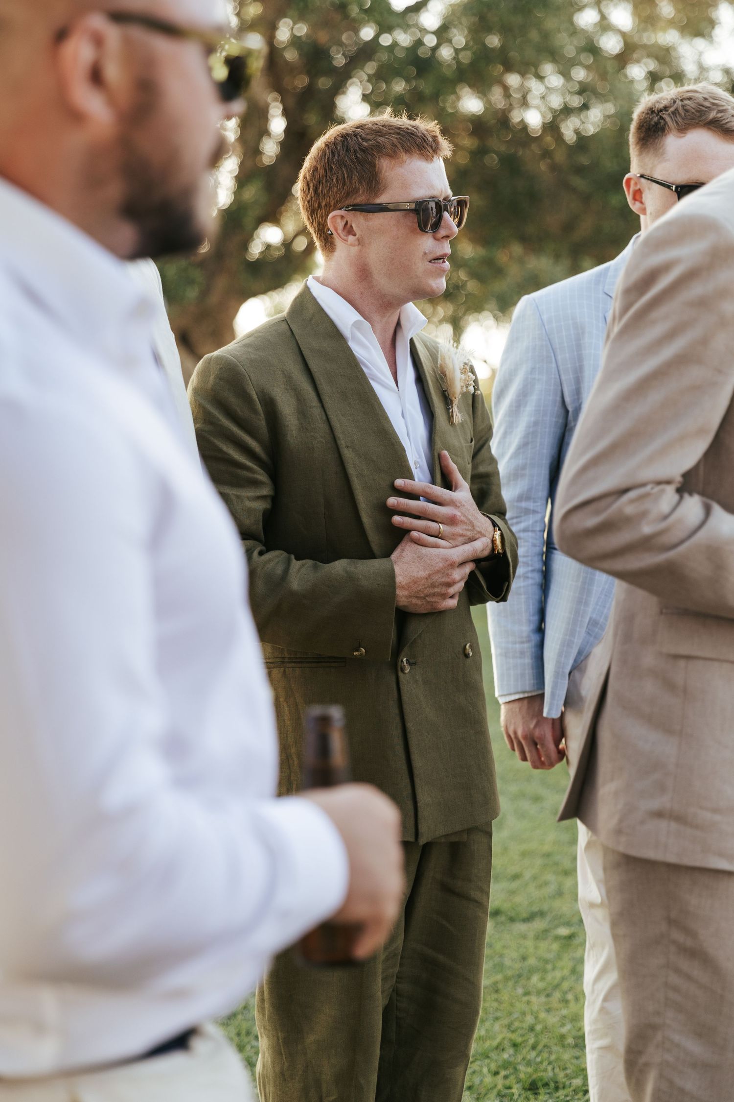 Groom in green double-breasted suit and sunglasses conversing with guests at outdoor wedding.