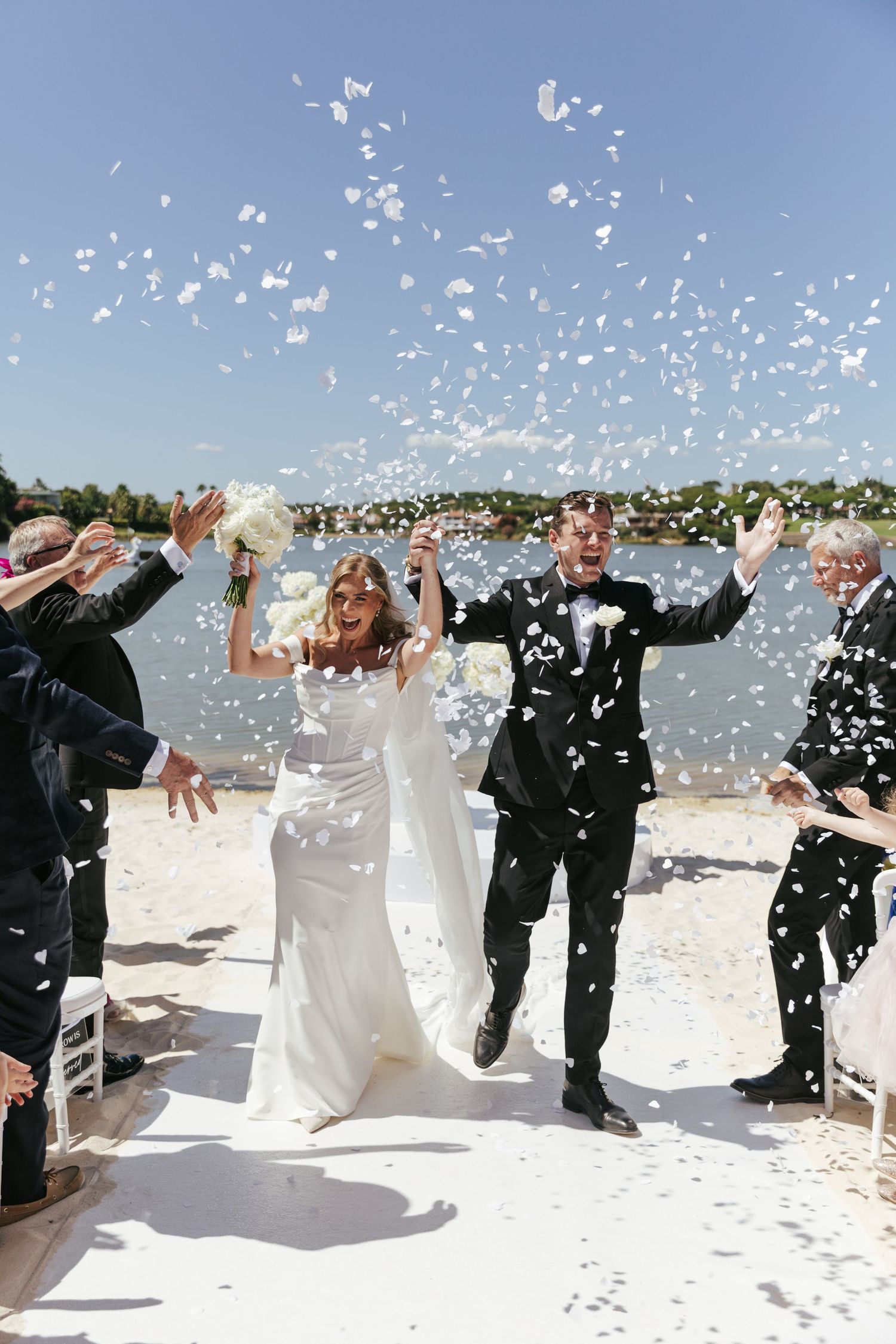 Joyful bride and groom celebrate on a beach aisle as guests toss white confetti on a sunny wedding day.