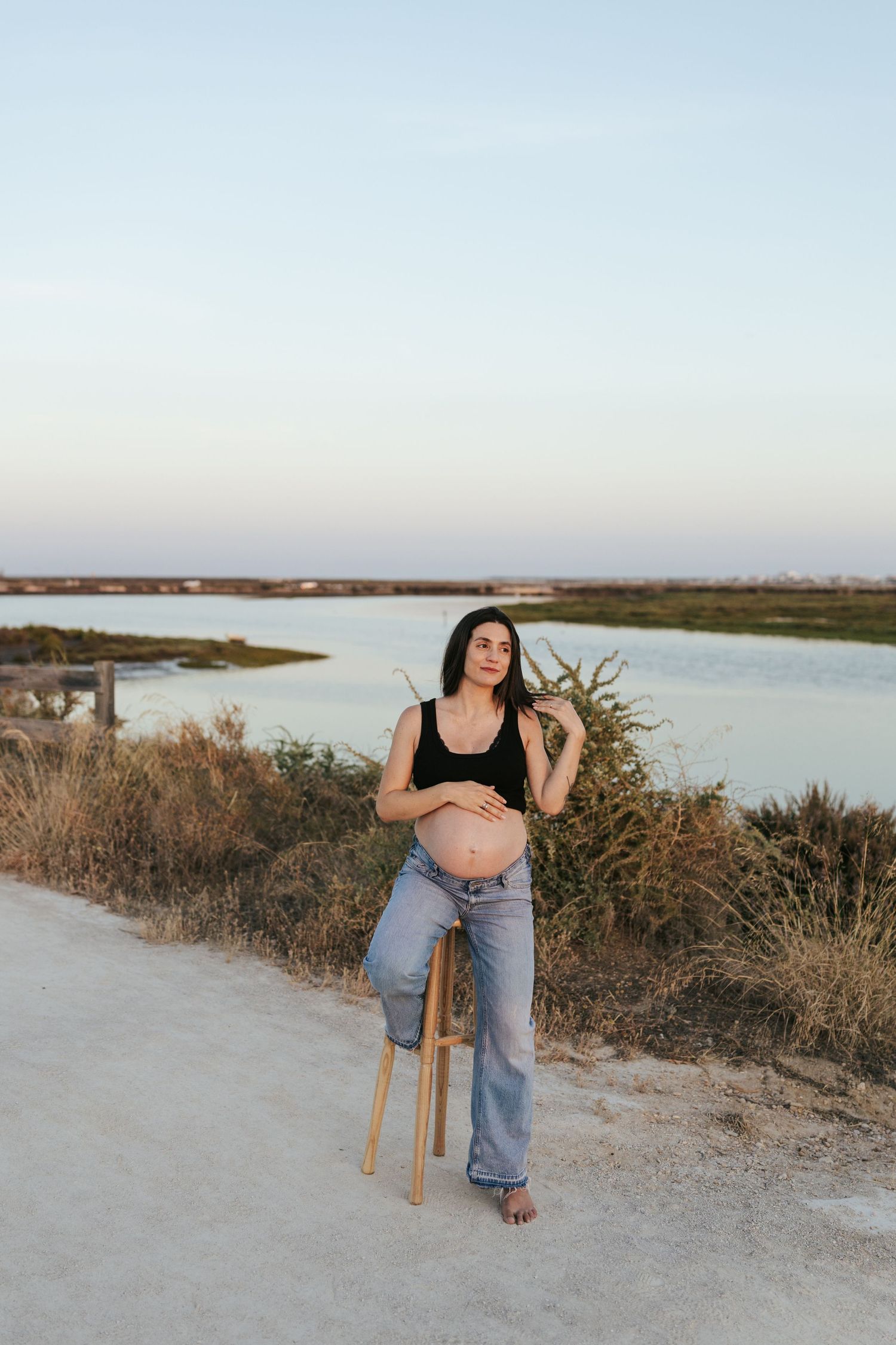 Pregnant woman in black crop top and jeans sitting on stool by a serene river at sunset.