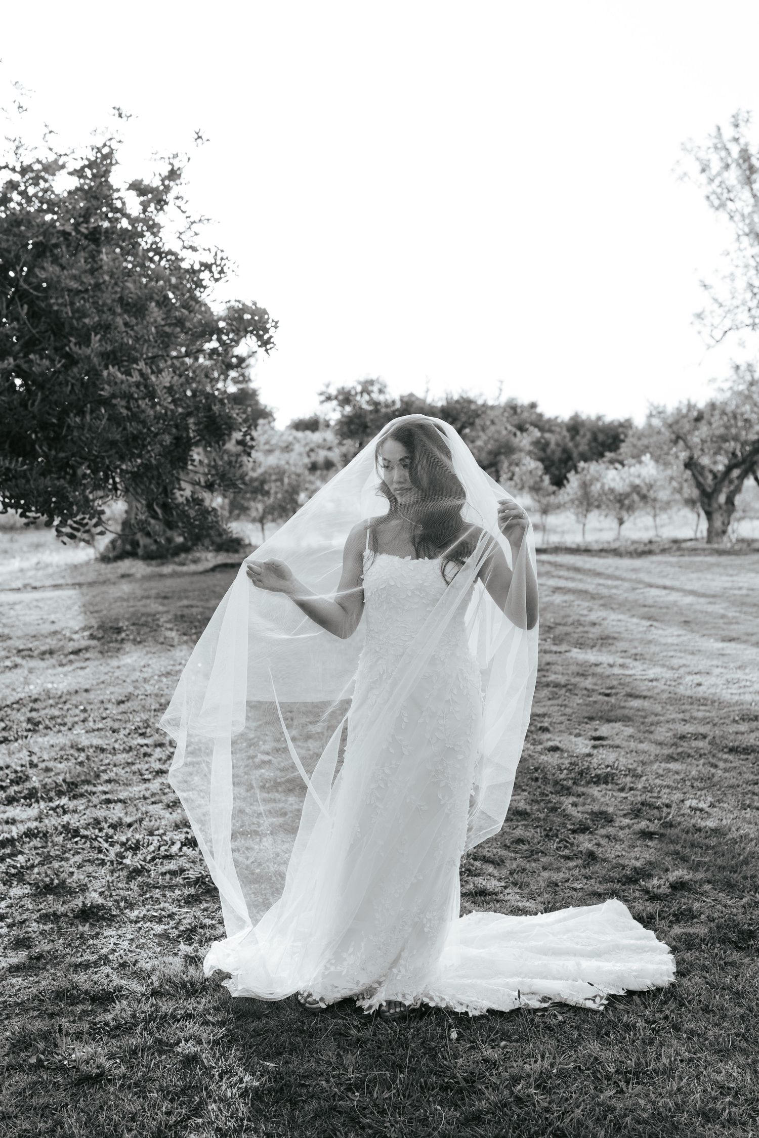 Bride in white gown lifting her veil outdoors in a black and white wedding photo.