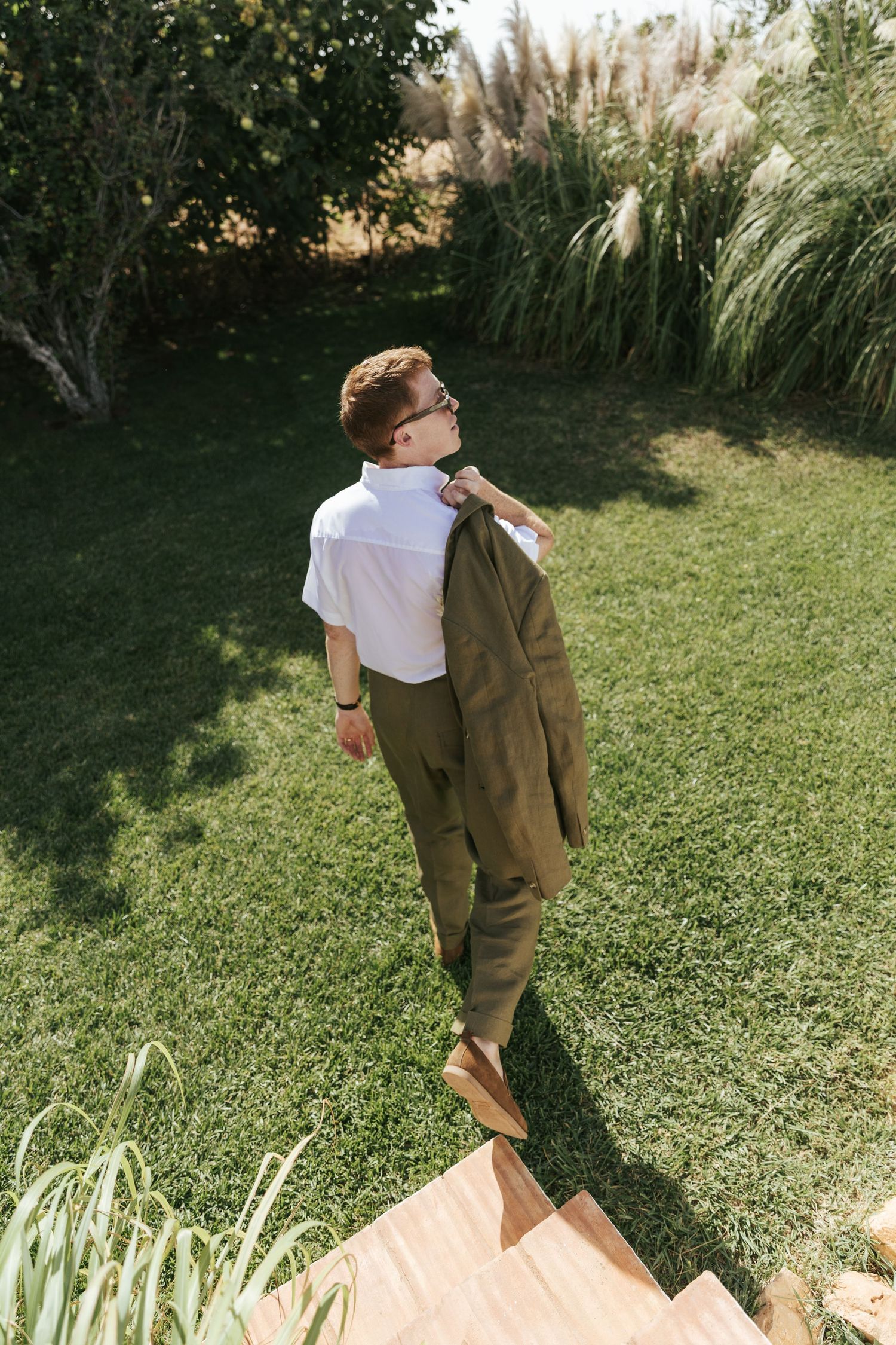 Young man in olive trousers and white shirt carries jacket over shoulder while standing on garden steps.