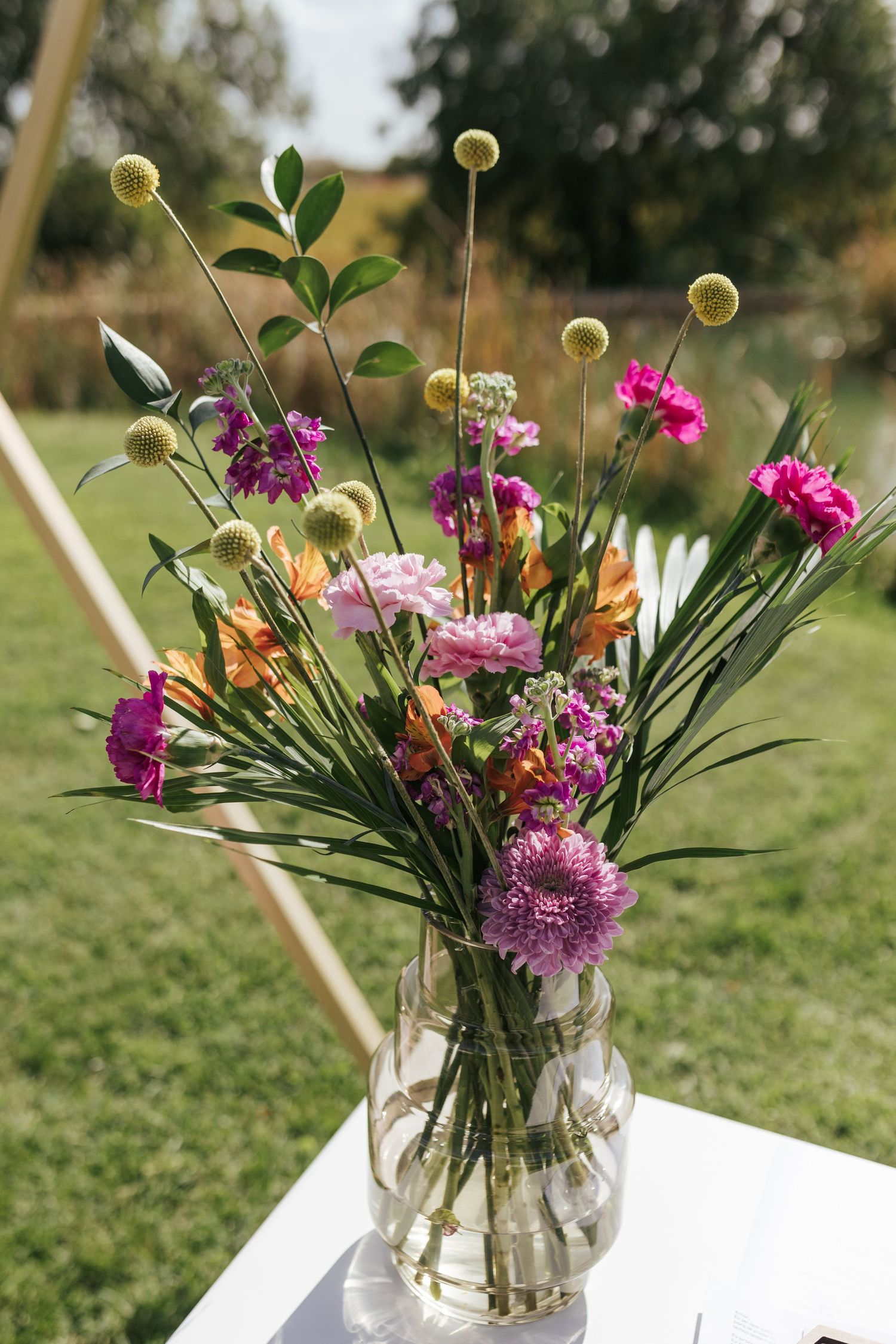 Colorful wedding bouquet with pink carnations, yellow craspedia, and mixed flowers in clear vase against garden backdrop.