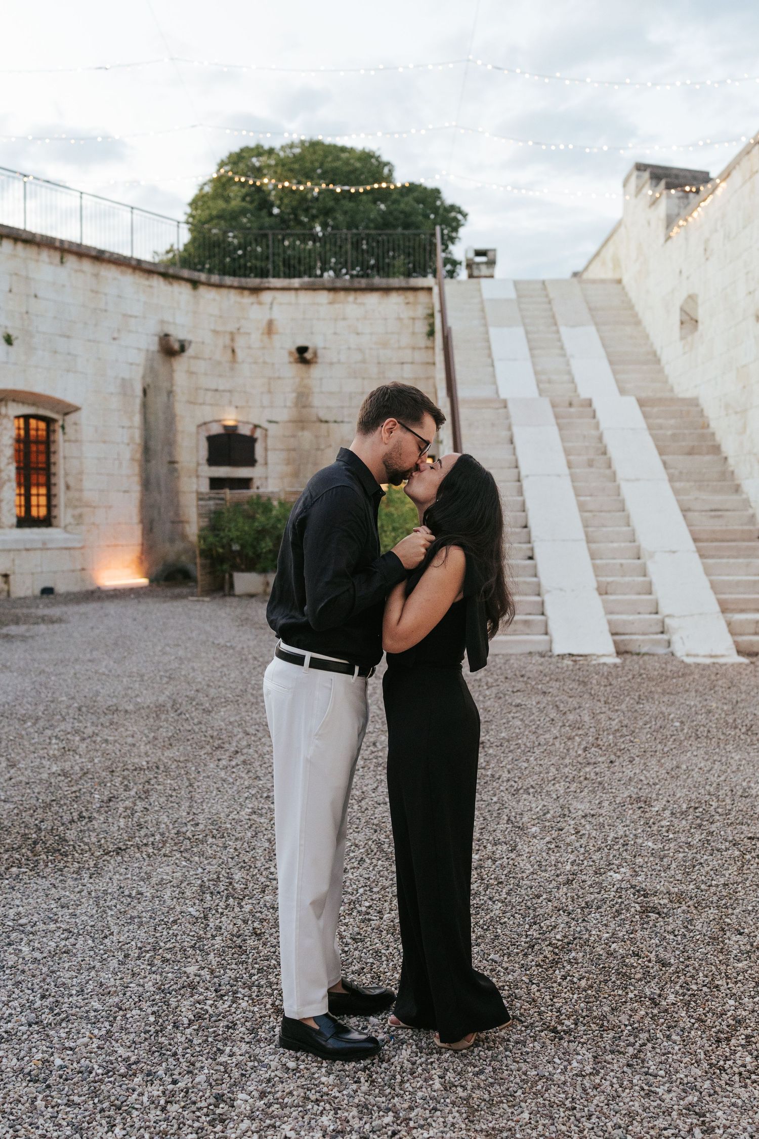 A romantic couple shares an intimate moment on a gravel courtyard with white stone walls and steps in the background.