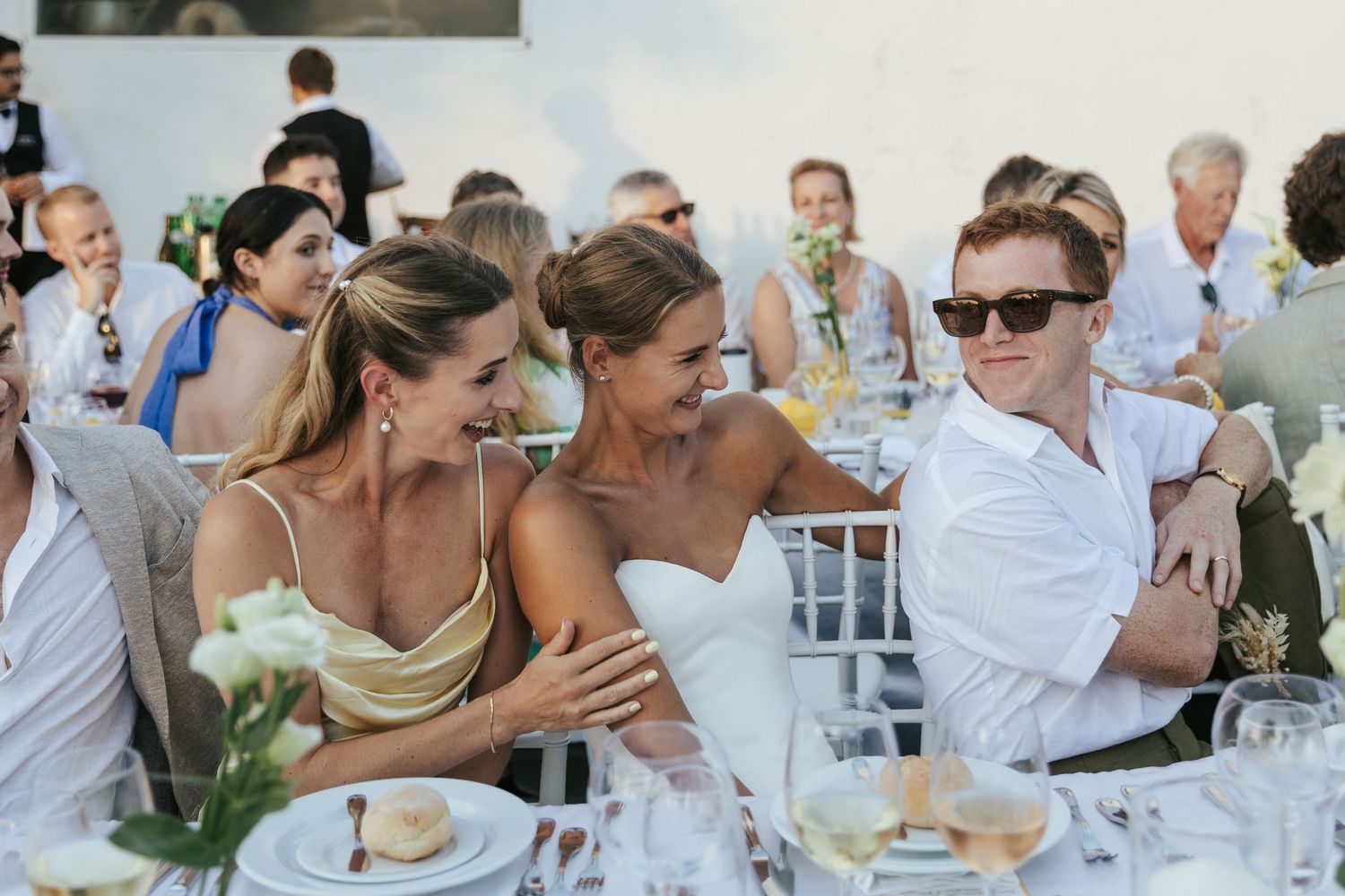 Three elegantly dressed guests laughing and socializing at an outdoor wedding reception dinner table.