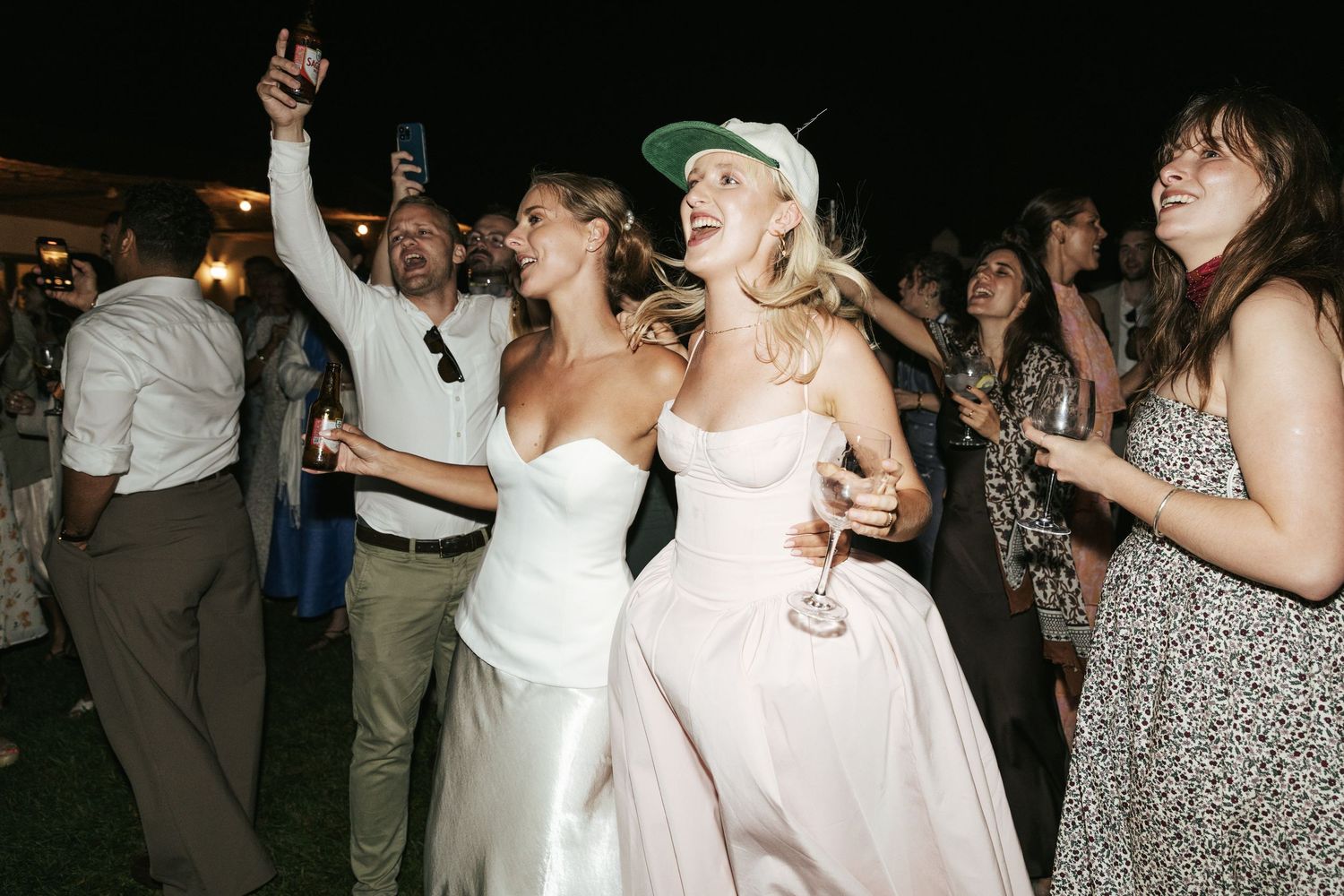 Two brides in white gowns dancing joyfully with guests at an outdoor wedding reception celebration.