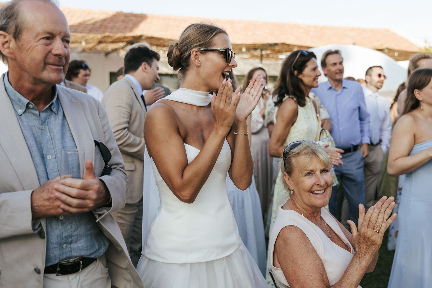 Bride in white dress clapping joyfully with wedding guests at an outdoor evening reception.