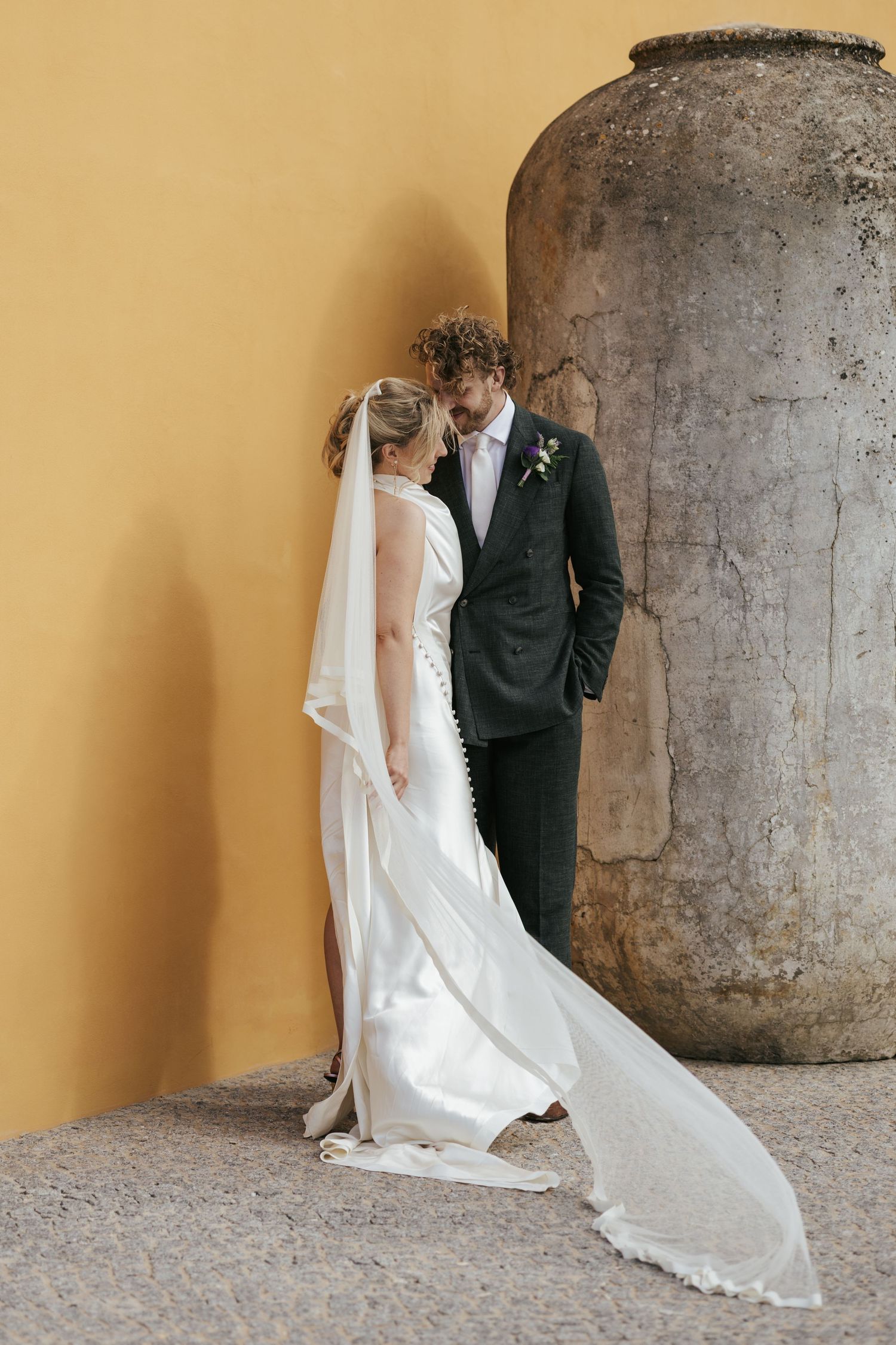 A couple dressed in wedding attire share an intimate moment against a yellow wall and stone column.