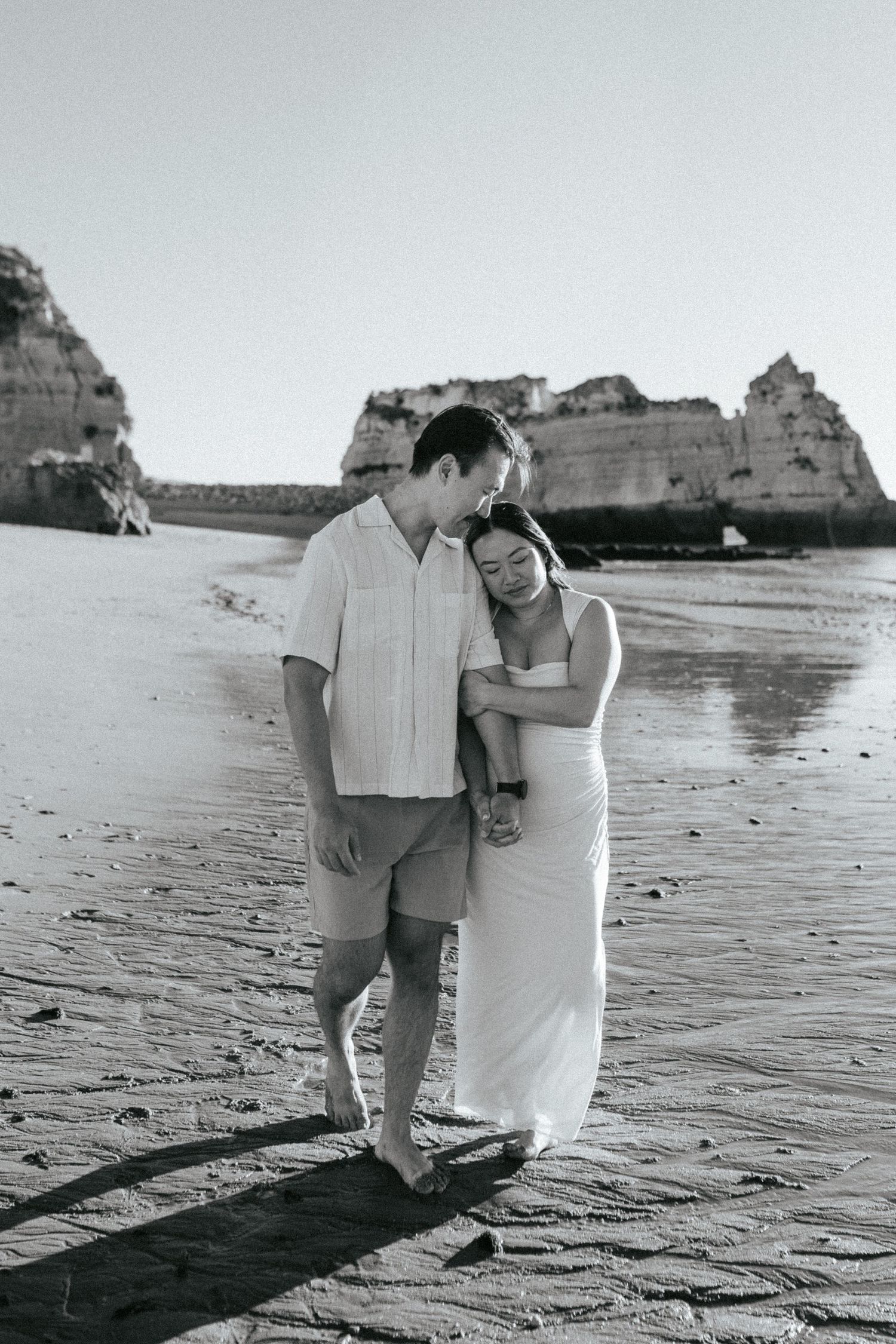 A romantic couple shares a tender moment on a beautiful beach with dramatic coastal cliffs in the background.