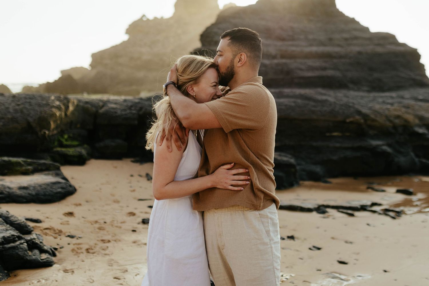 Two people embrace in a tender moment on a rocky beach at sunset with dramatic cliffs in the background.