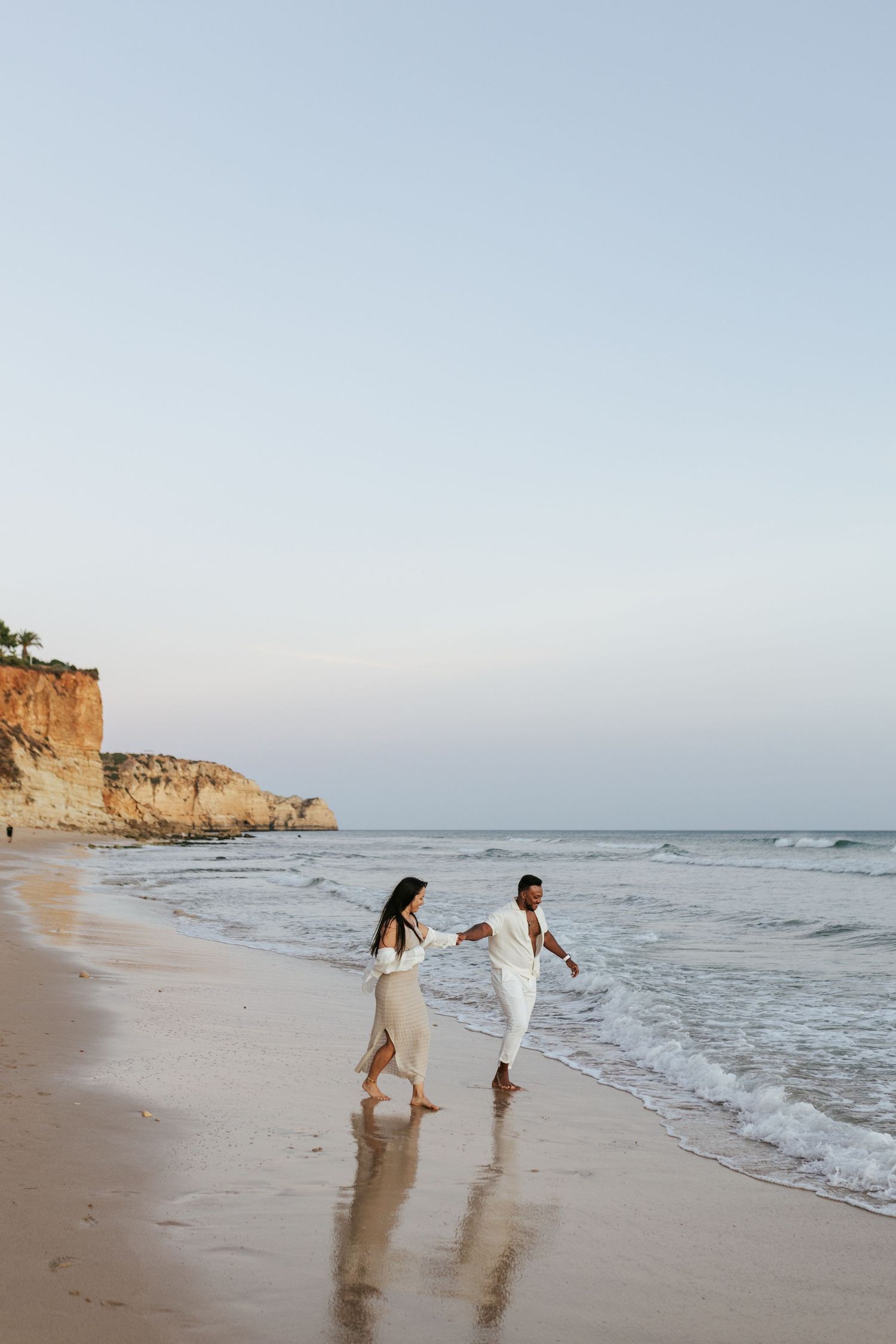 A couple in light colored outfits walks playfully along the shoreline near coastal cliffs. Praia do Porto de Mós, Lagos, Algarve, Portugal.