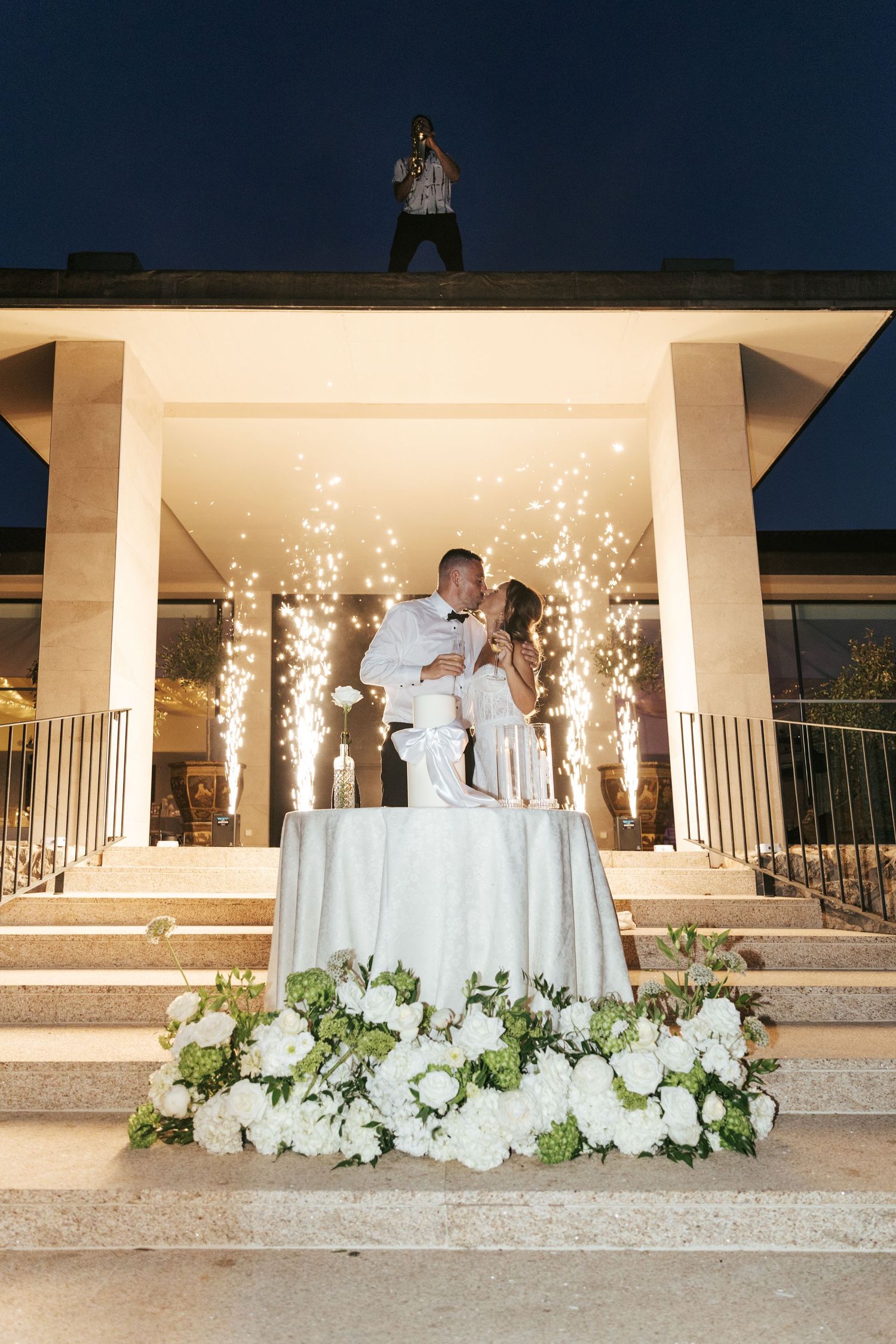 Couple sharing a kiss at wedding cake table on illuminated steps with white floral arrangements and sparkler backdrop at night.