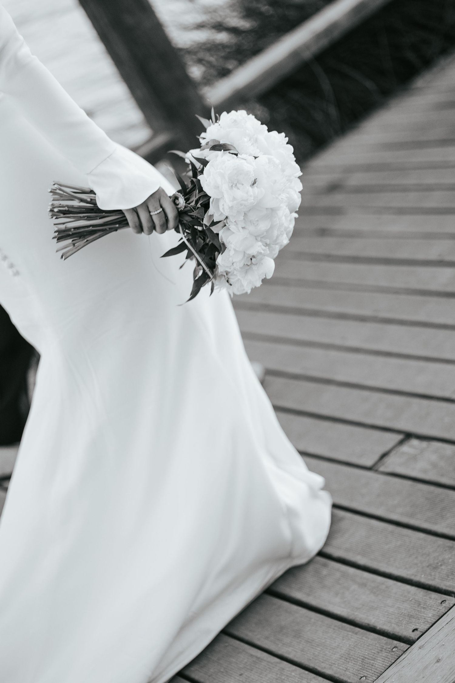 Black and white photo of a bride in a white gown holding a floral bouquet on a wooden dock.
