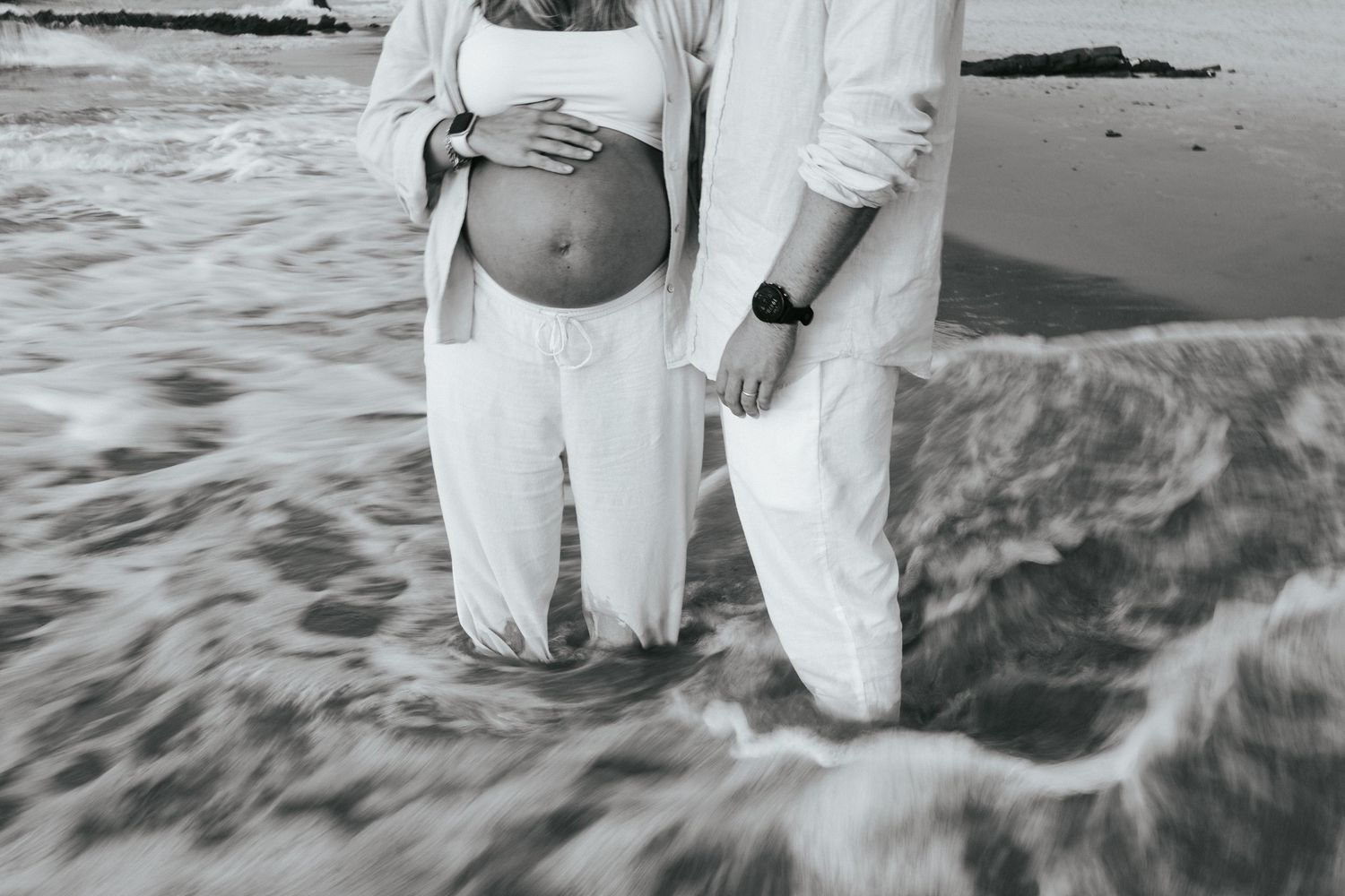A couple in white outfits walks along a scenic beach shoreline with waves crashing at their feet.