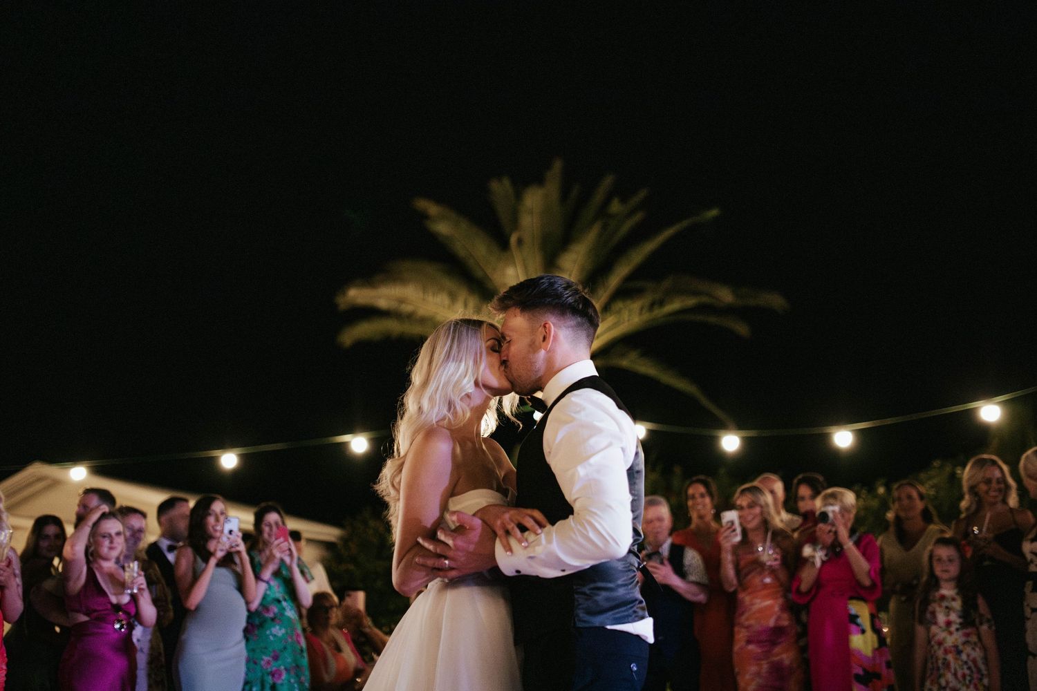 A crowd of wedding guests dance together under string lights during an outdoor evening reception celebration.