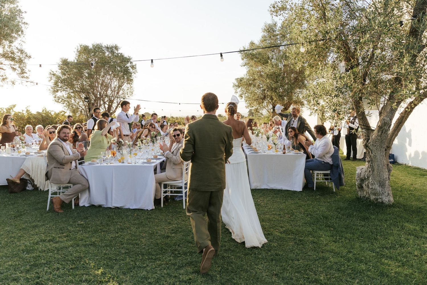 Bride and groom walk toward guests seated at outdoor wedding reception tables surrounded by olive trees.