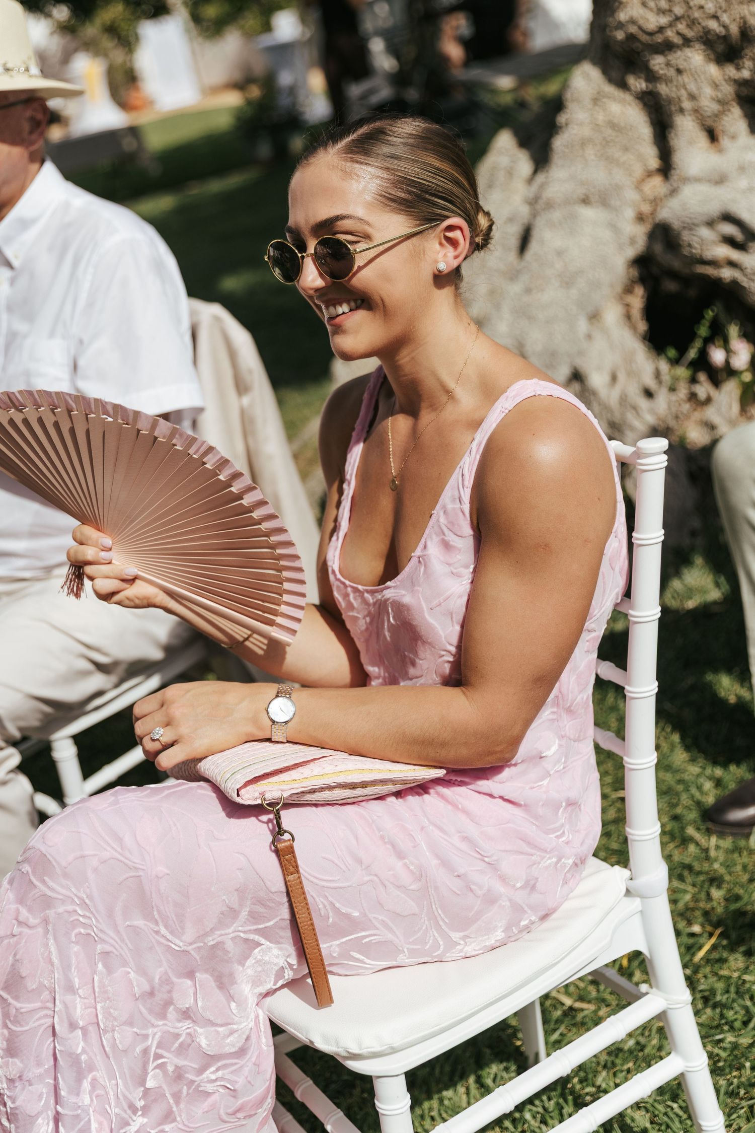 Smiling woman in pink dress and sunglasses holding wooden fan at outdoor summer wedding.