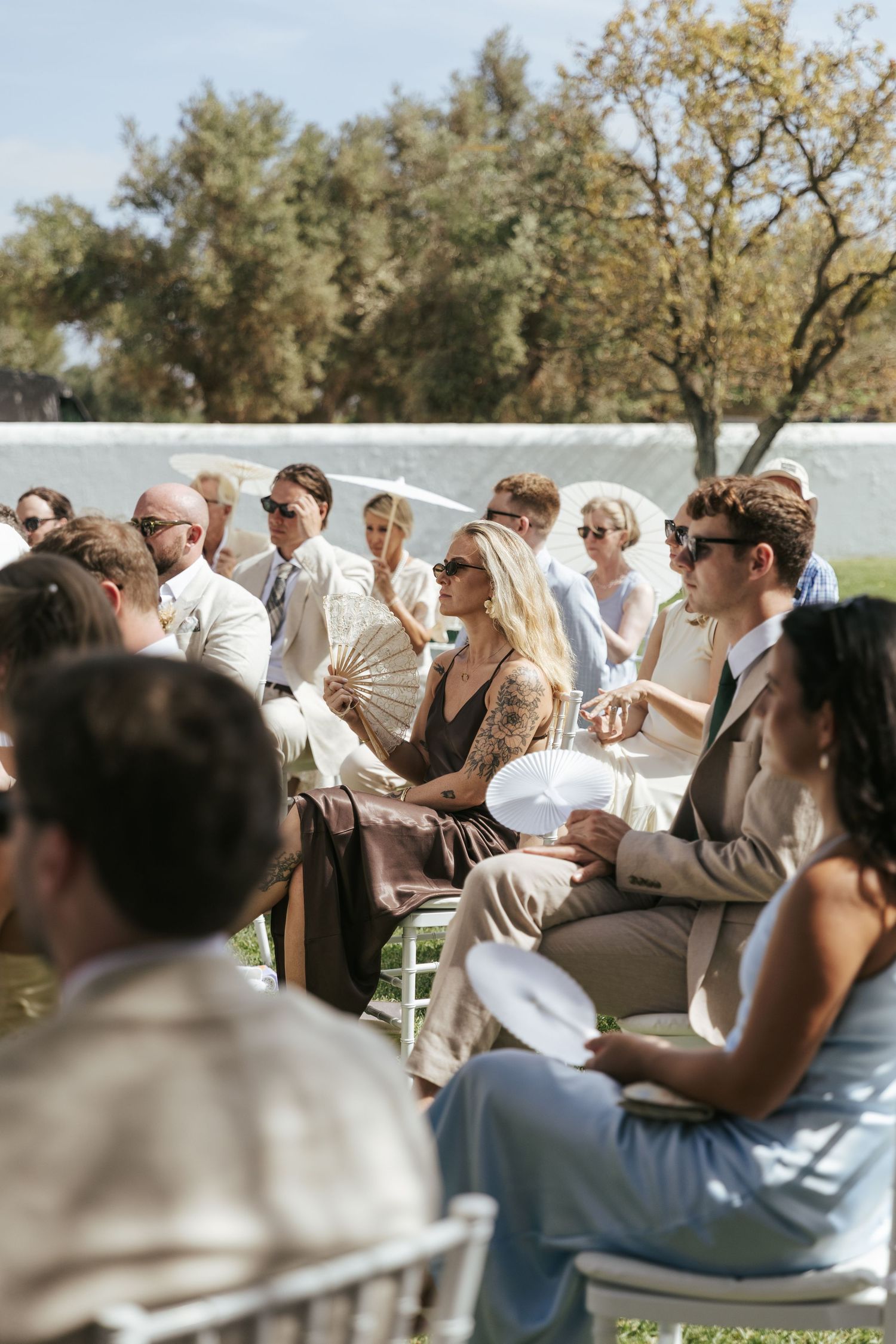 Outdoor wedding guests seated on white chairs, fanning themselves in the sun, dressed in elegant summer attire.