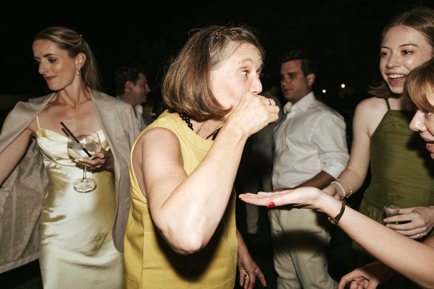 Woman in yellow dress laughing while eating at lively outdoor nighttime wedding reception party.