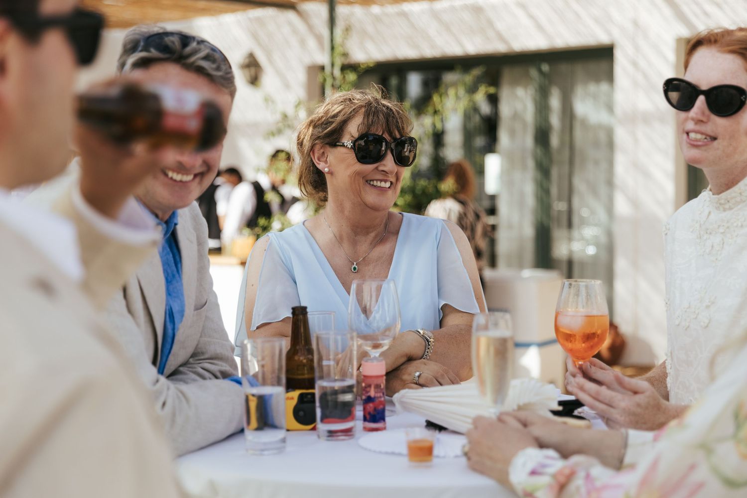 Guests laughing and socializing at an outdoor wedding reception table with drinks.