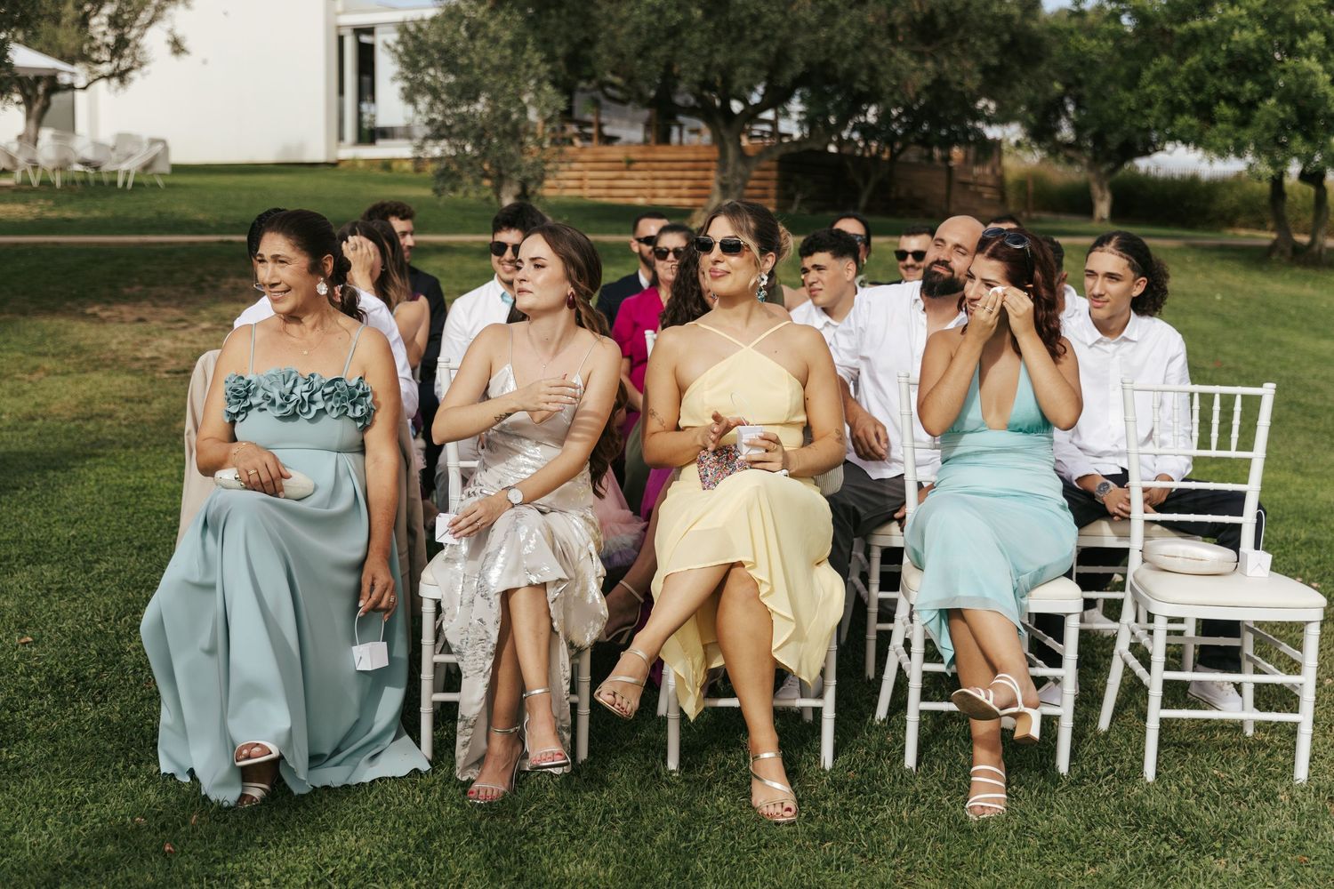 A group of bridesmaids in pastel dresses laughing and reacting emotionally during an outdoor wedding ceremony.
