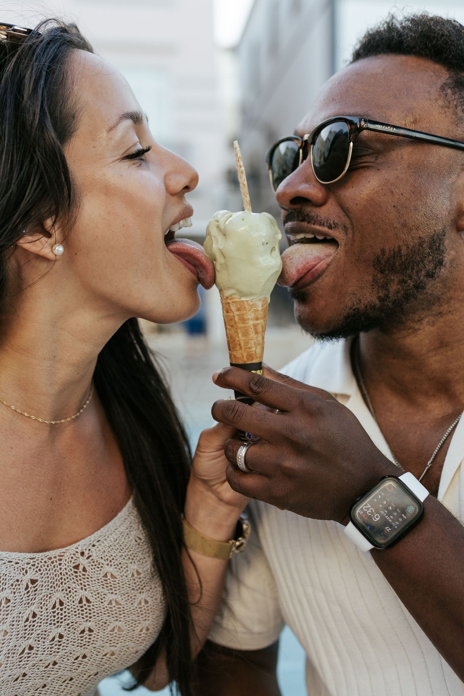 A playful couple sharing an ice cream cone outdoors while sticking their tongues out to lick it.