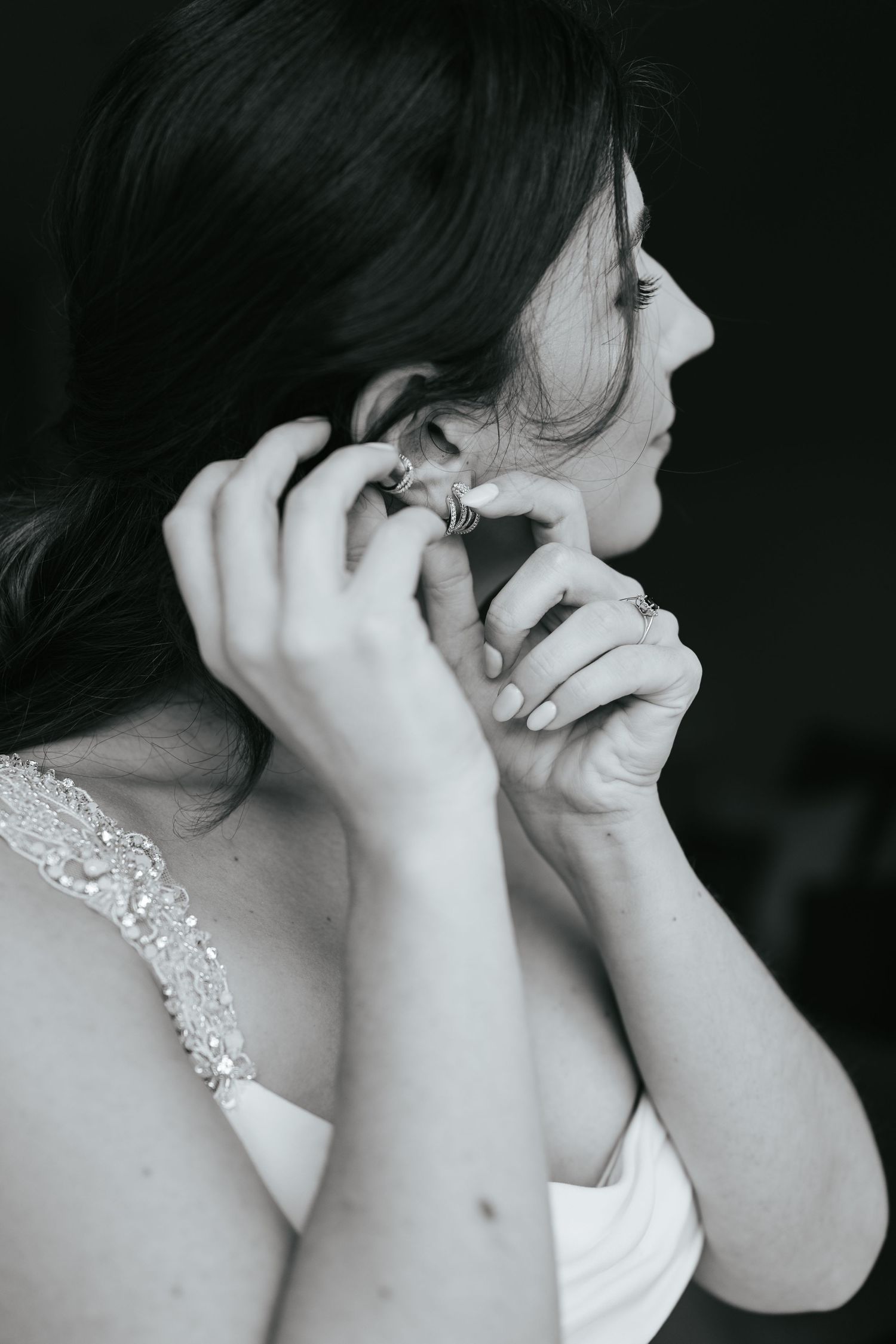 Black and white photo showing someone putting on an earring while wearing a sleeveless white dress with beaded straps.