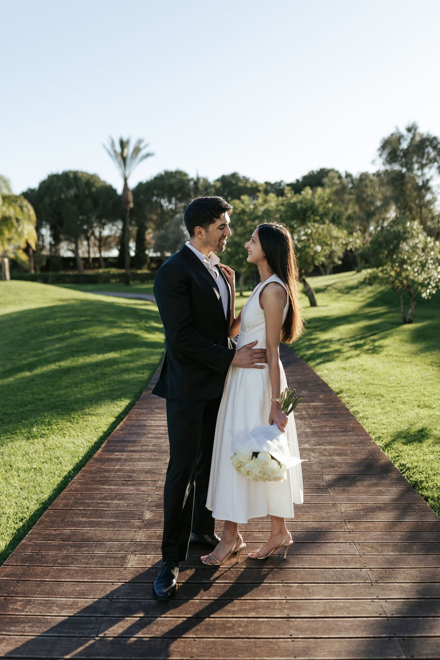 A couple stands together on a wooden boardwalk surrounded by lush green landscaping on a sunny afternoon.