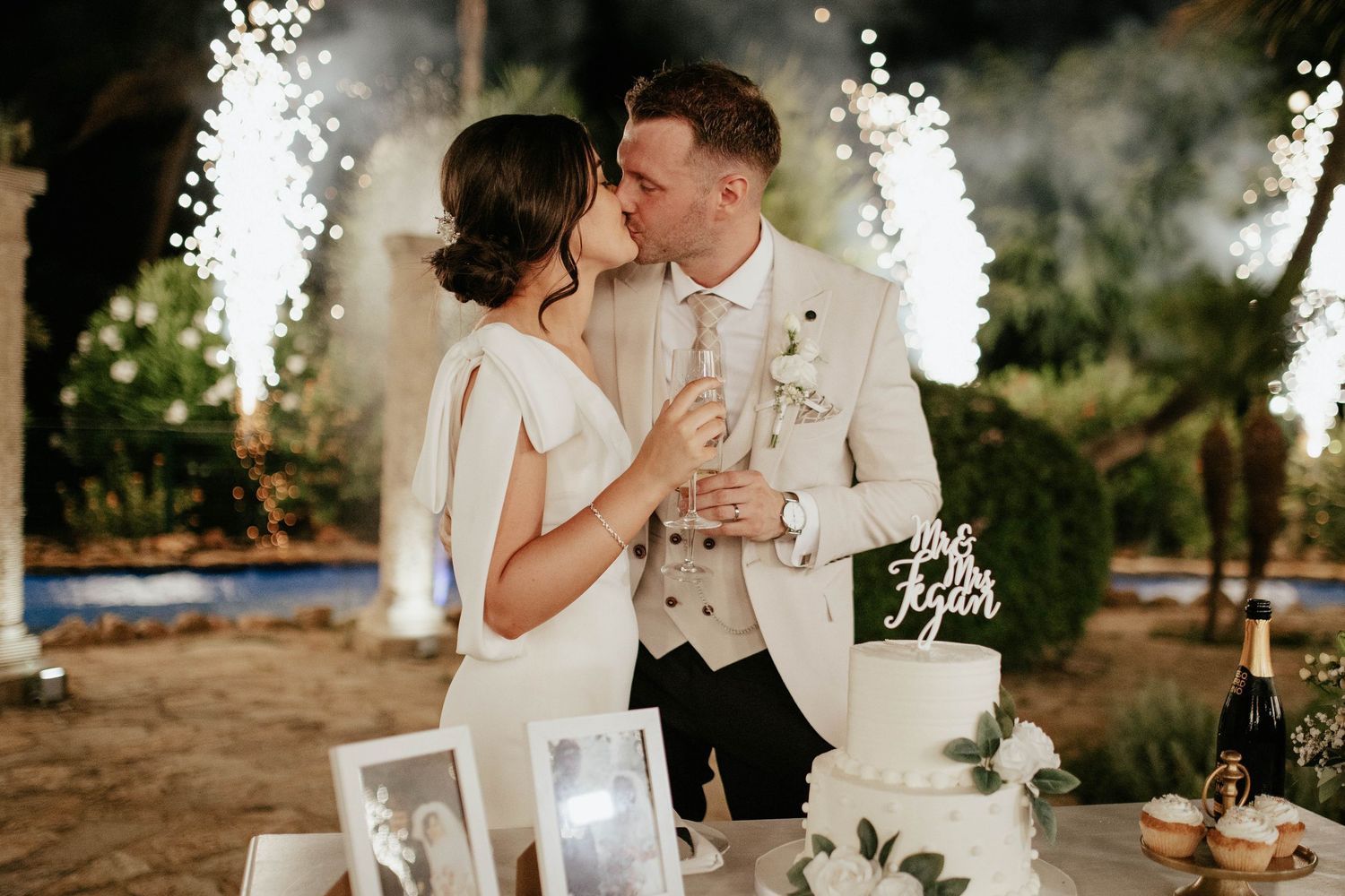 Newlyweds share romantic kiss at outdoor wedding reception with sparklers and elegant white cake display.