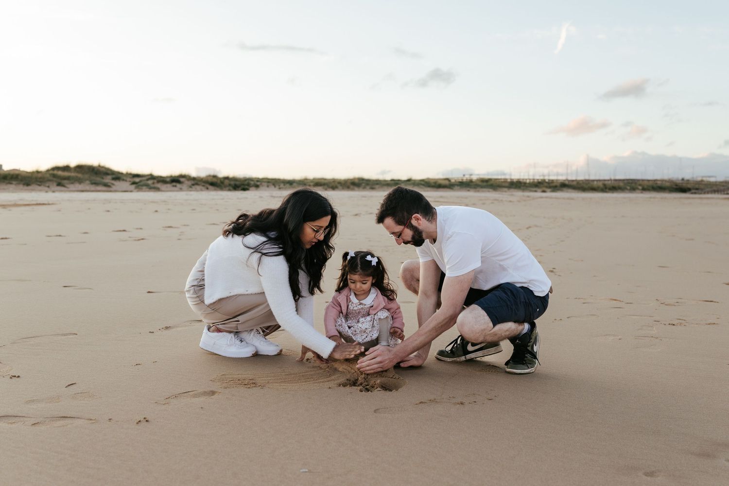 Three people crouched together on a sandy beach during sunset exploring the sand.