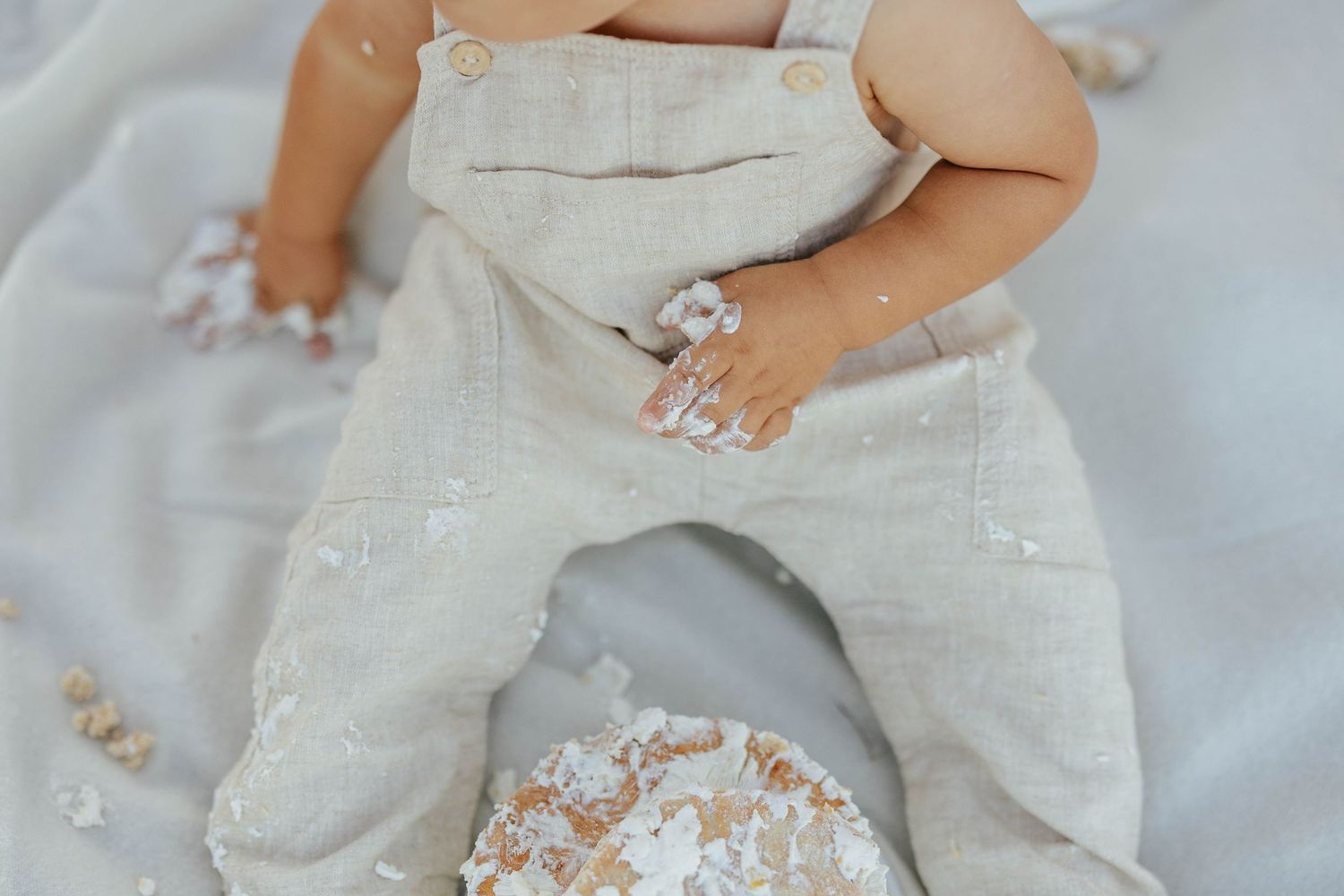 Baby wearing light gray linen overalls covered in flour while playing on messy kitchen counter.