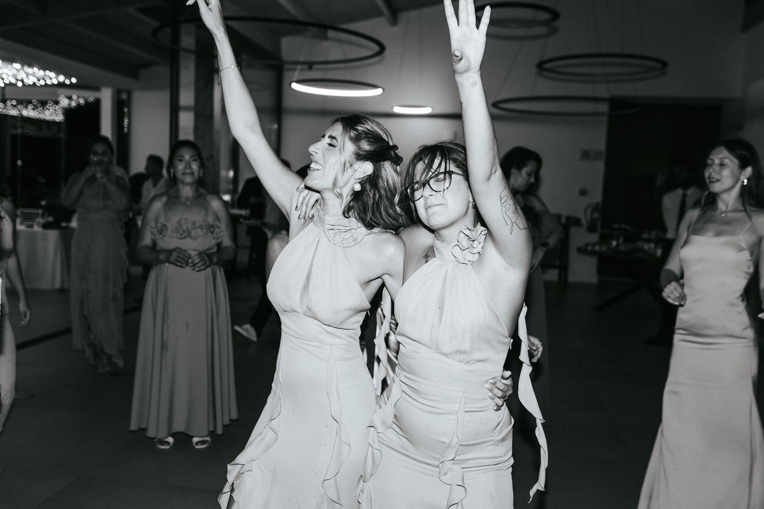 Two people in formal dresses dance with raised hands at what appears to be a wedding reception celebration.