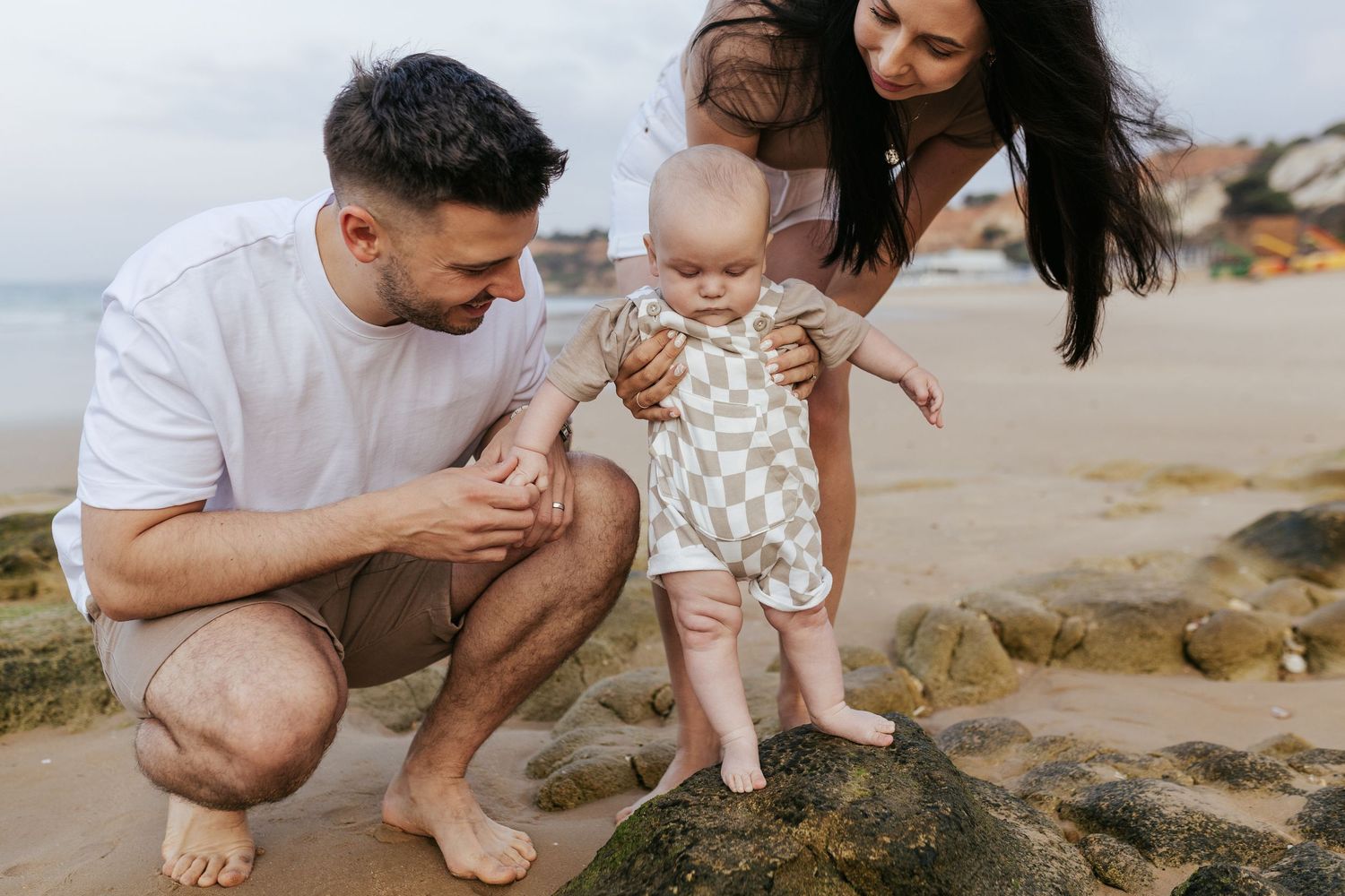 A loving family enjoys a tender moment together on a sunny beach near rocky coastal formations.