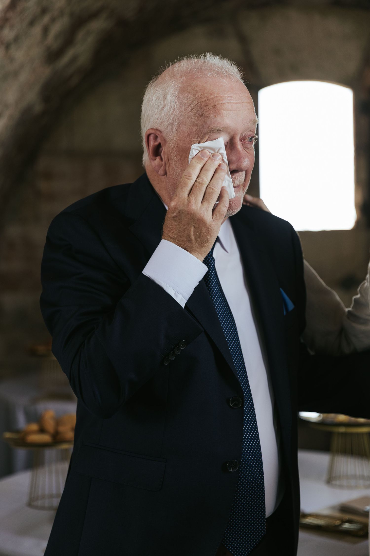 Person in dark suit wiping face with white handkerchief in dimly lit room.